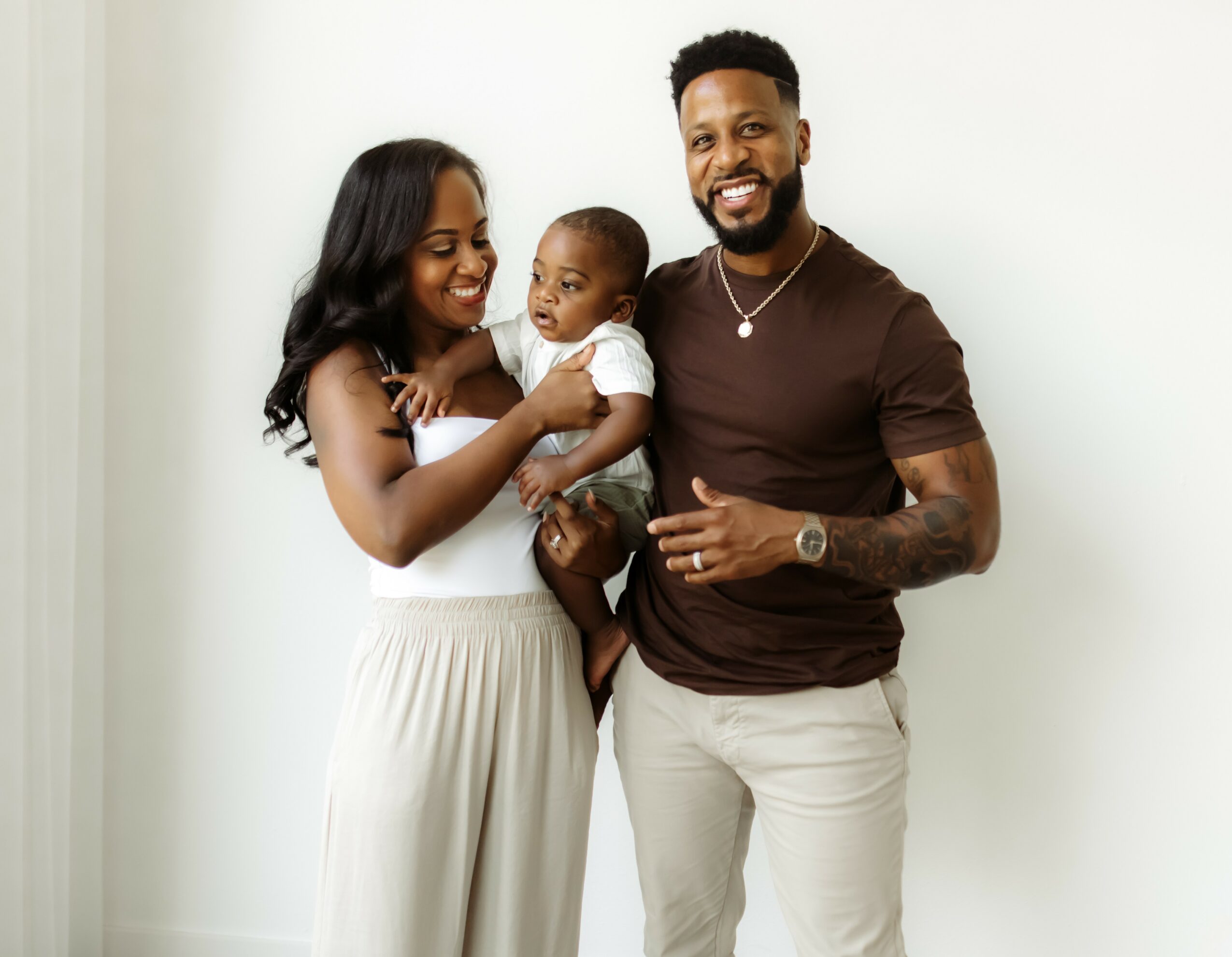 Family of three smiling, woman holding baby, man standing beside them, against a plain white background.