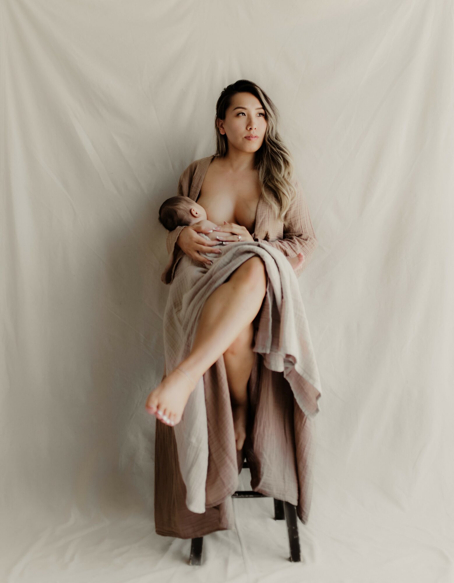 Woman sitting on a chair, holding a baby, wrapped in a blanket, with a neutral background.