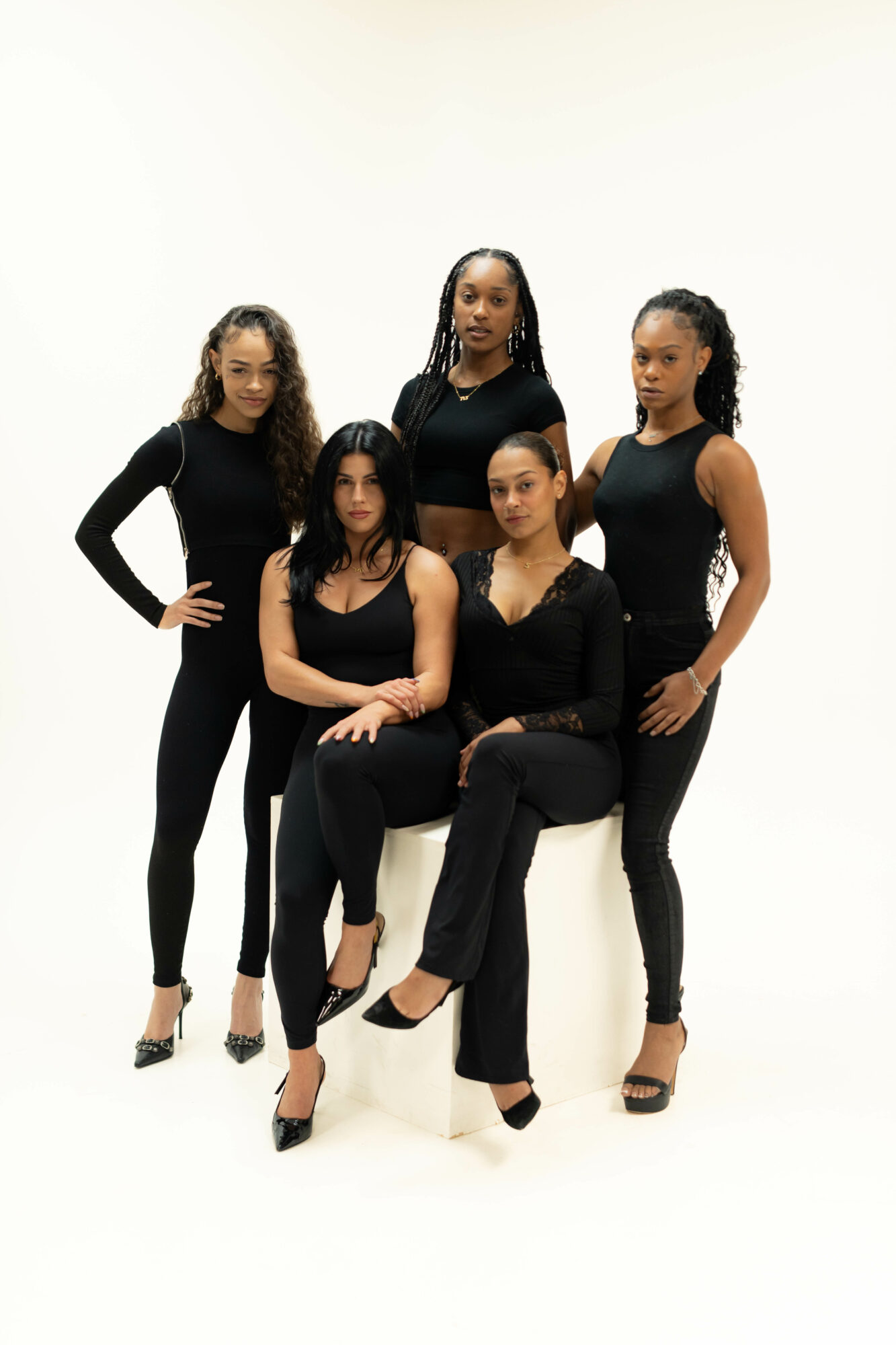 Five women in black outfits pose against a white background, some standing and some sitting, with confident expressions.