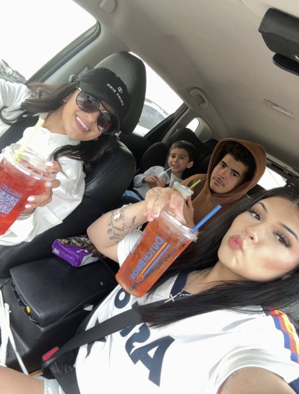 Four people sitting in a car, two women in the front holding drinks, two children in the back seat, smiling and posing.
