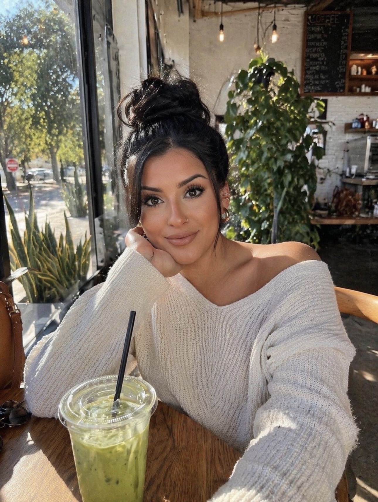 Young woman with dark hair in a bun, wearing a white off-shoulder sweater, sitting at a table with a green drink, smiling.