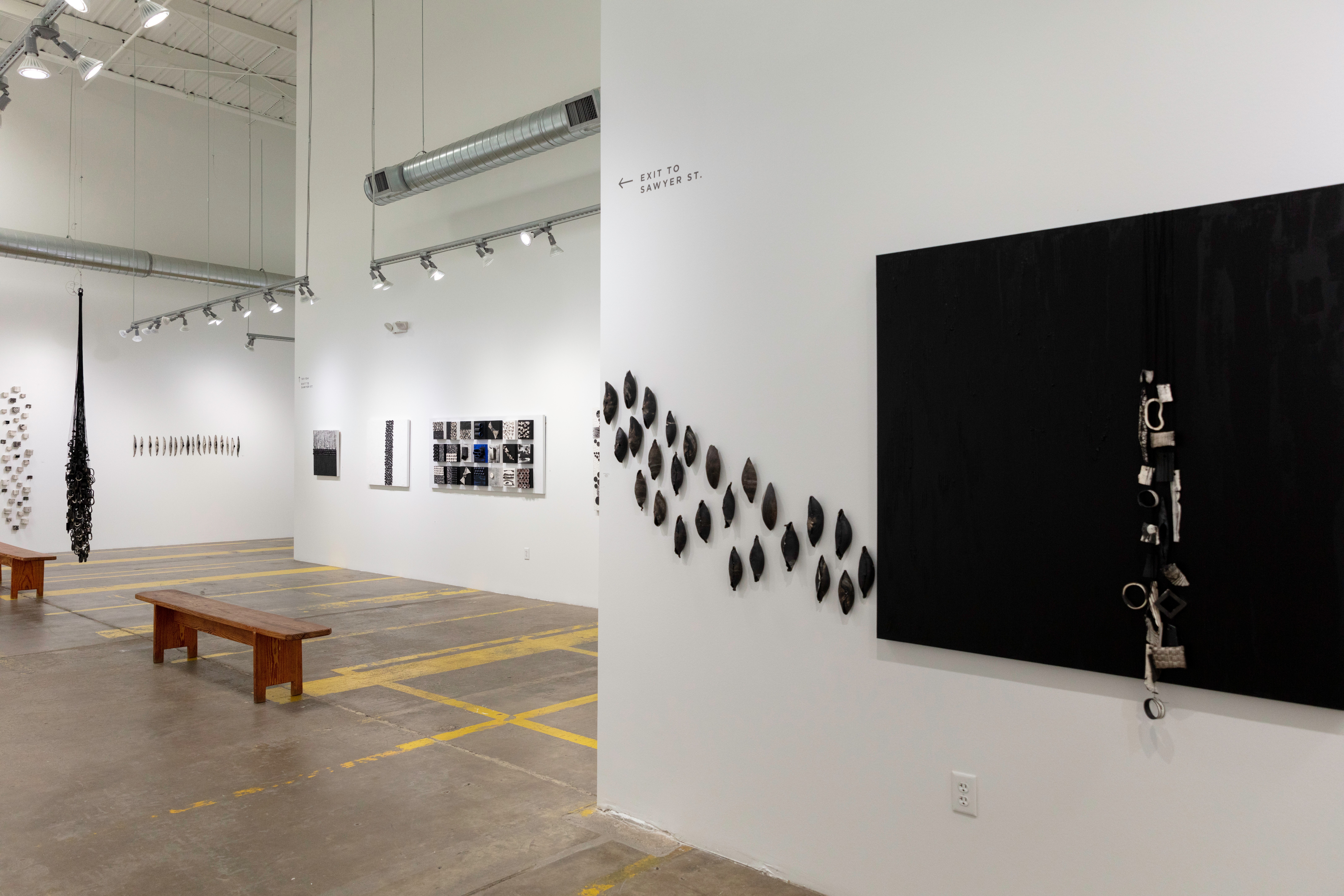 Art gallery with white walls, black and white artwork, wooden benches, and a high ceiling with lighting fixtures.