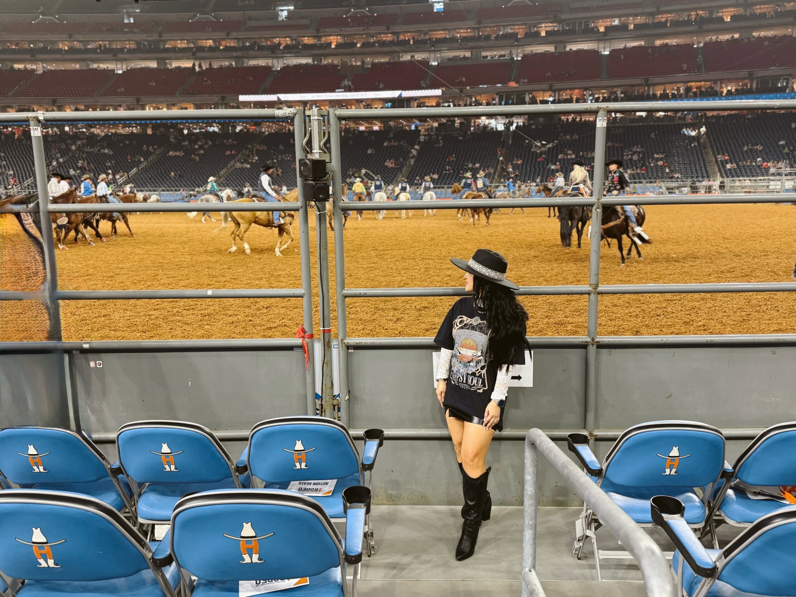 Person wearing a hat and black boots watching a horse race from a stadium seating area.