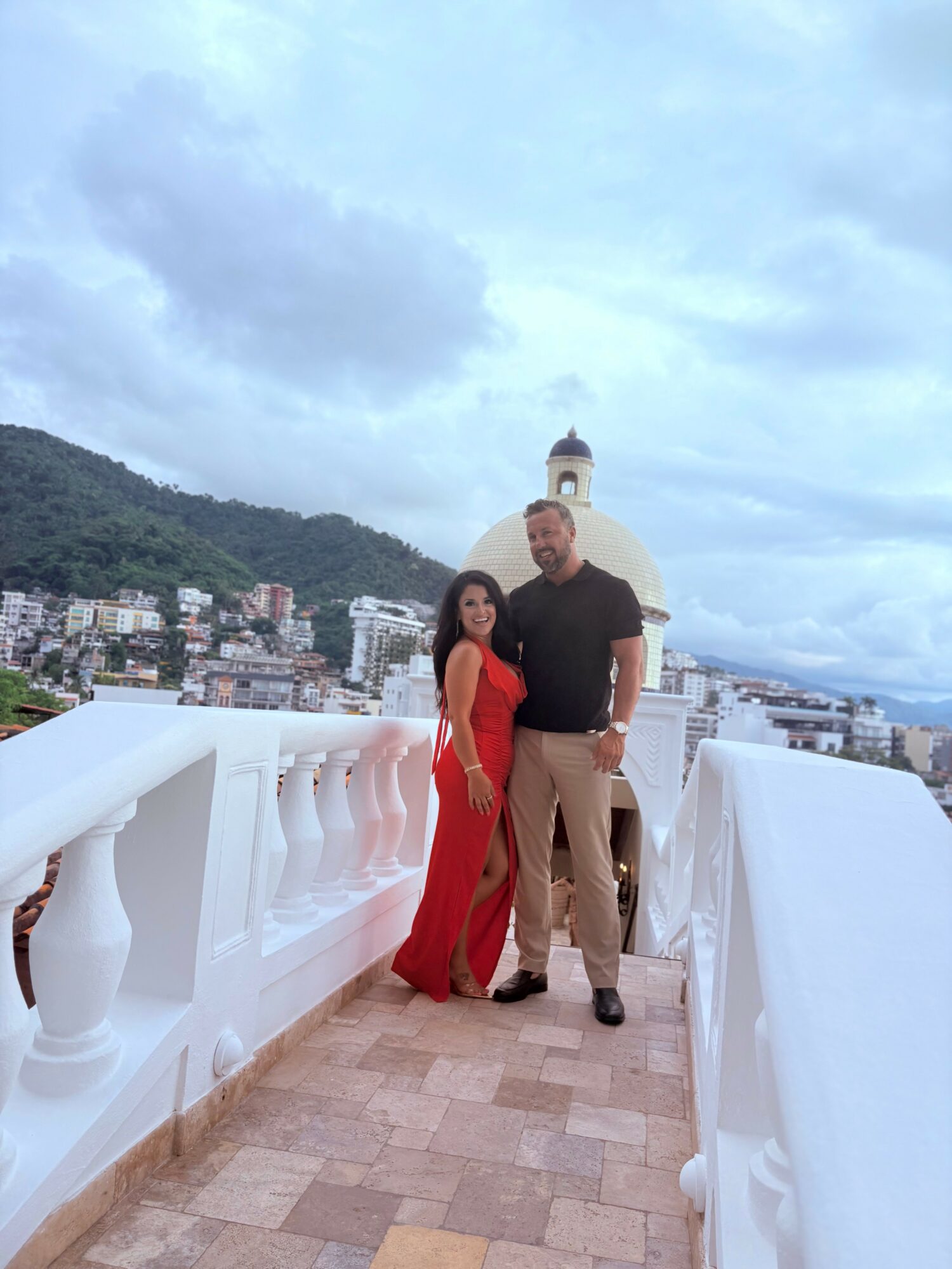 A man and woman stand on a white balcony with a cityscape and a dome in the background, under cloudy sky.