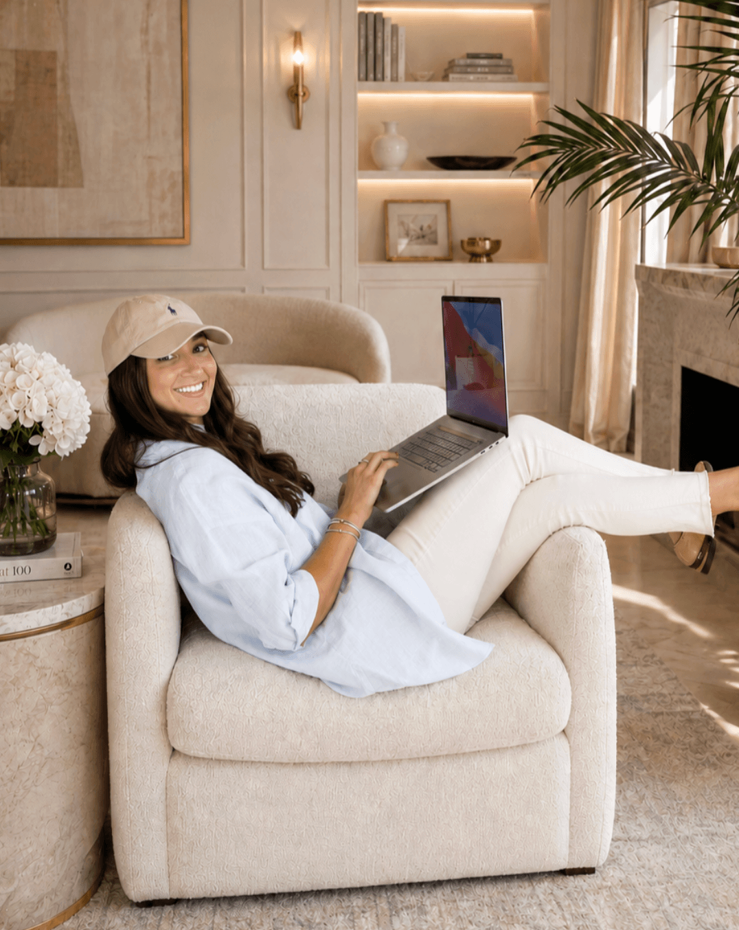 Woman sitting on a cream armchair with a laptop, smiling, in a well-lit living room with bookshelves and plants.