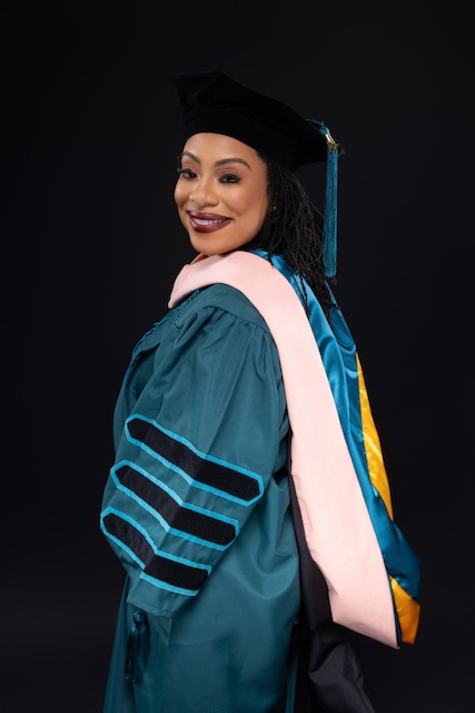 Woman in graduation gown and cap smiling, facing slightly to the side, against a dark background.