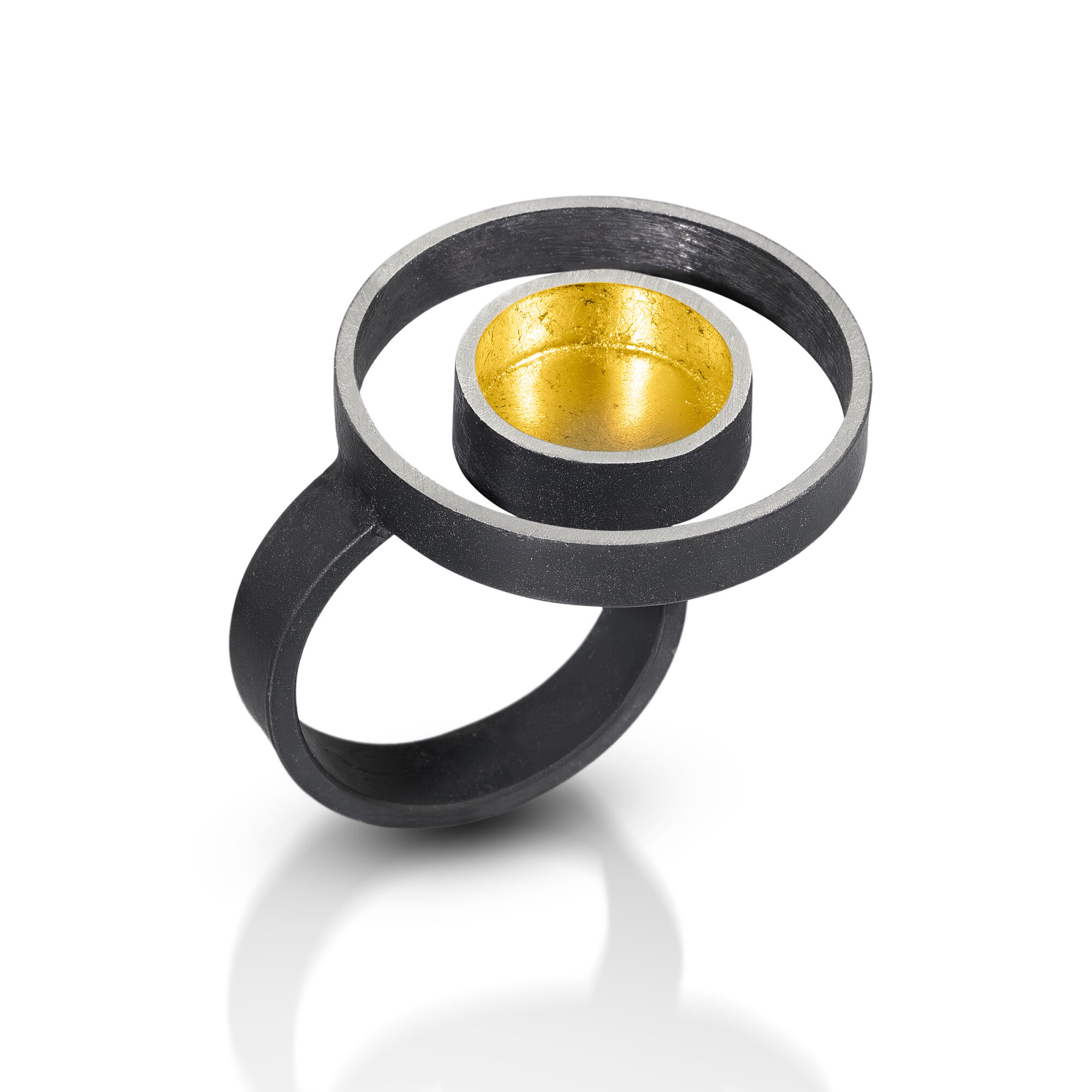 Ring with black outer structure and gold inner circle, connected by a black band, casting a shadow on white background.