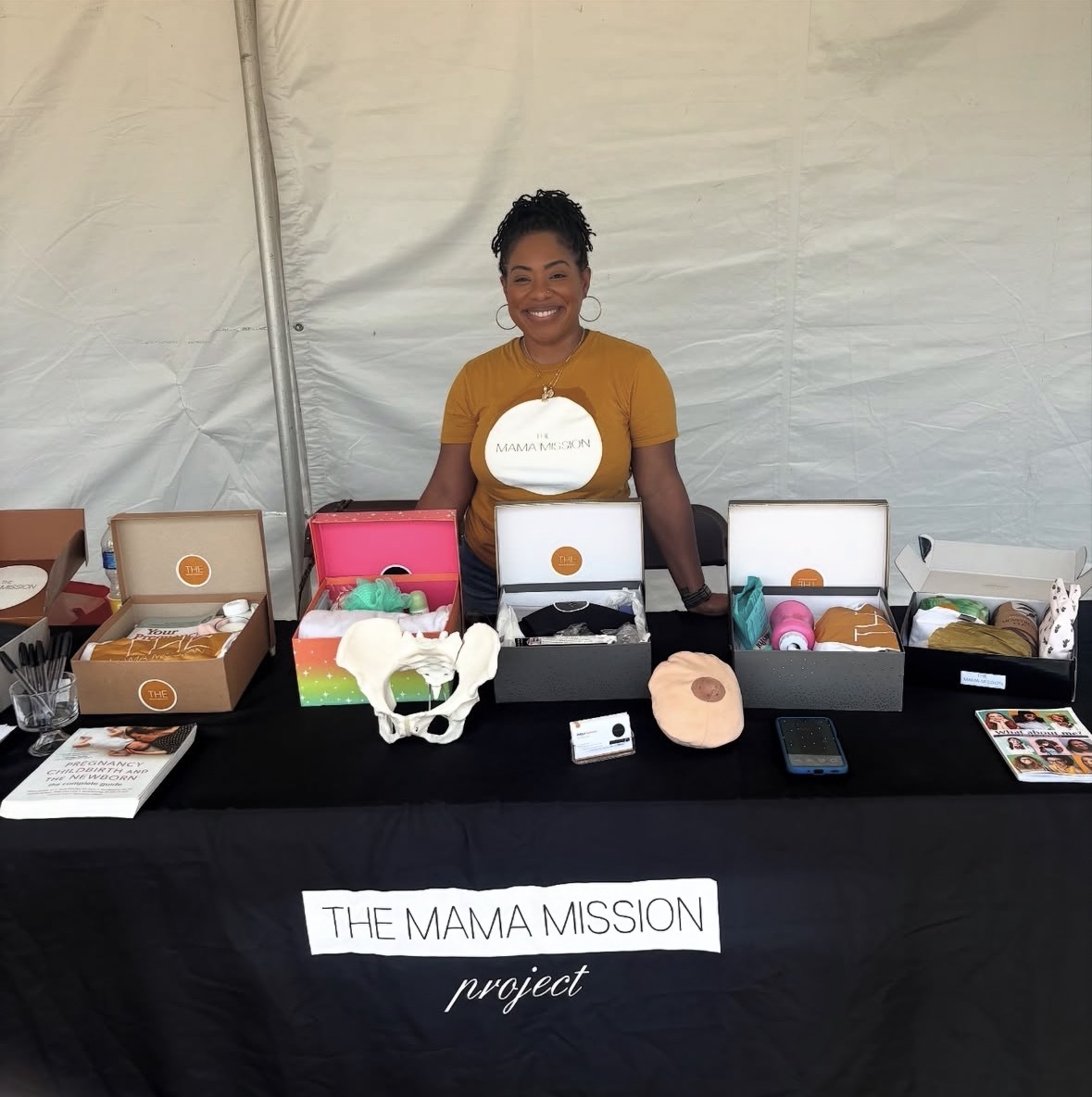 Smiling woman standing behind a table with various products, under a tent, promoting The Mama Mission project.