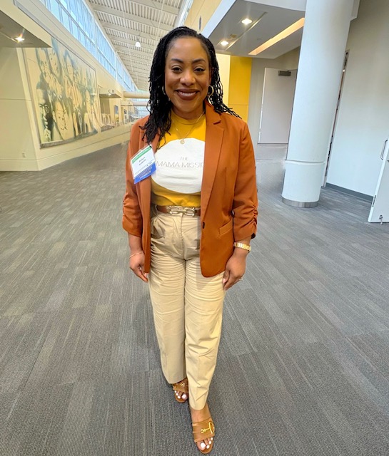 Woman standing indoors, smiling, wearing a brown blazer, yellow top, beige pants, and open-toe shoes.