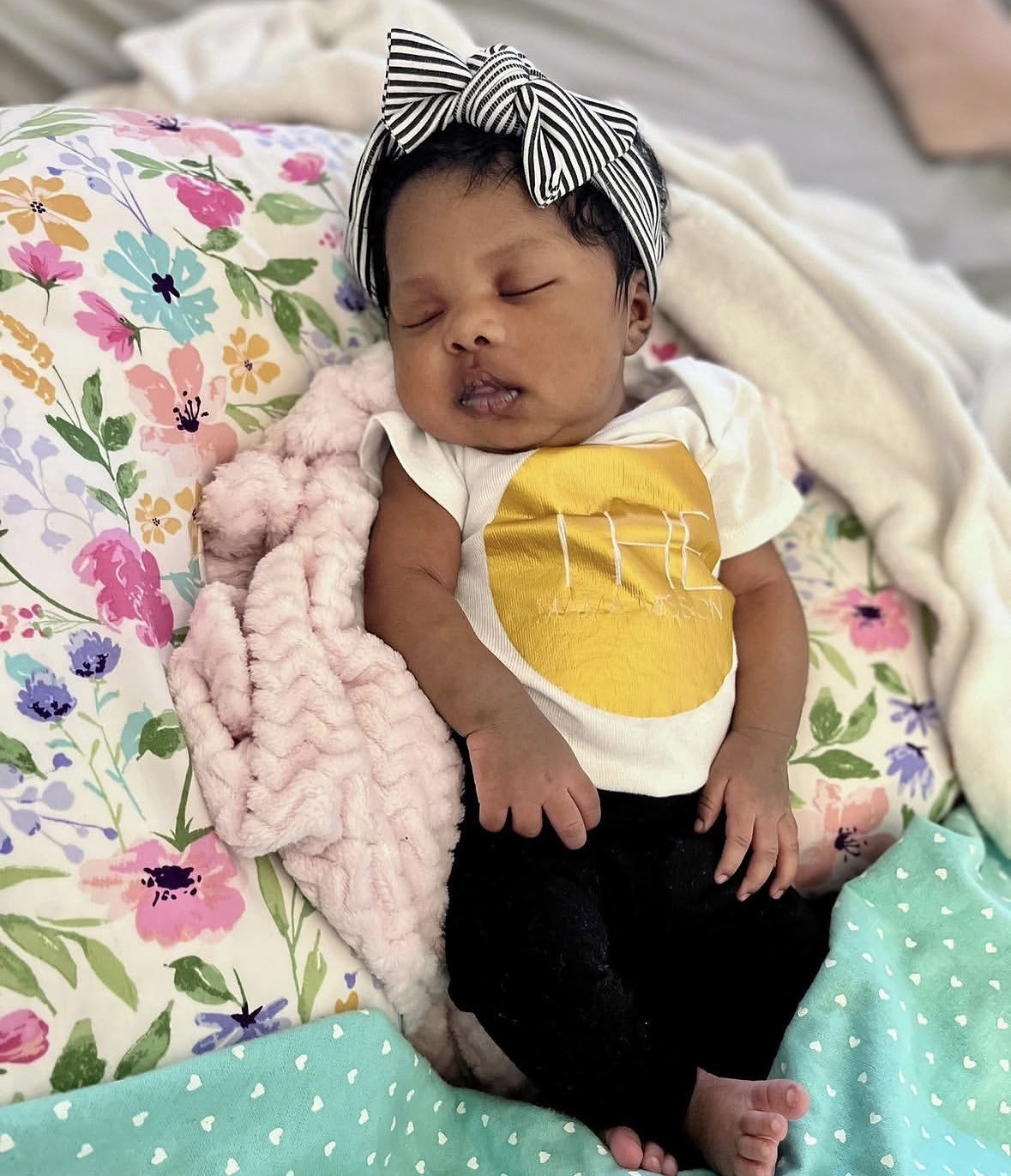 Baby sleeping on floral pillow with headband, pink blanket, and colorful bedding, wearing a yellow and white shirt and black pants.