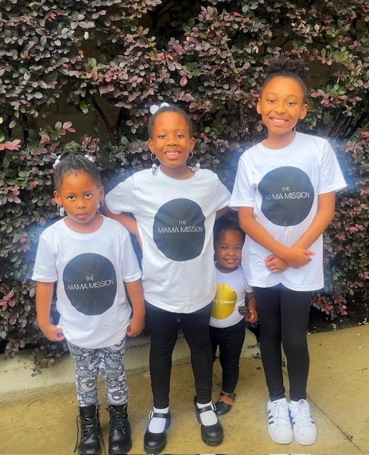Four children standing outdoors in front of flowering bushes, smiling, wearing white T-shirts with a black circle and text, with varied pants and shoes.