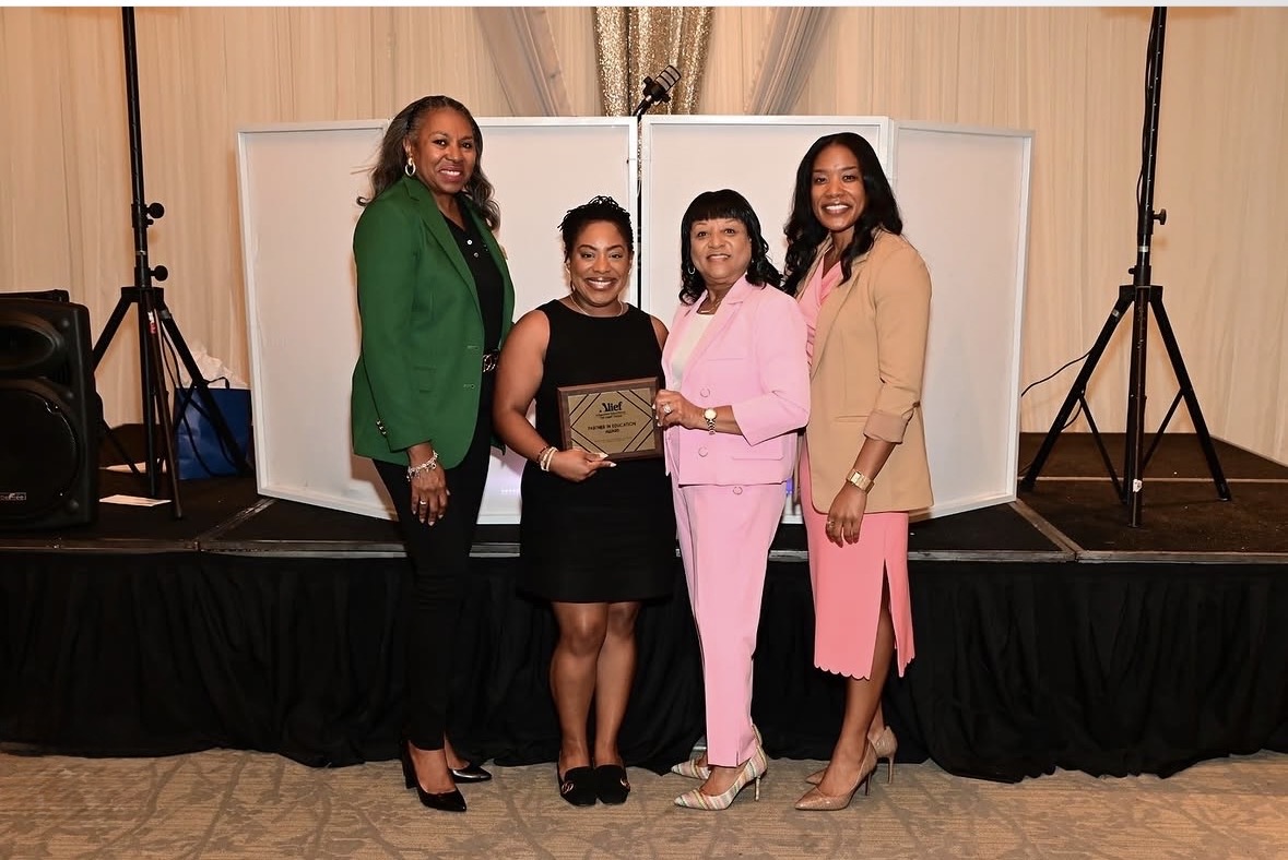 Four women standing on a stage, one holding a plaque, in front of white panels, smiling, in formal attire.