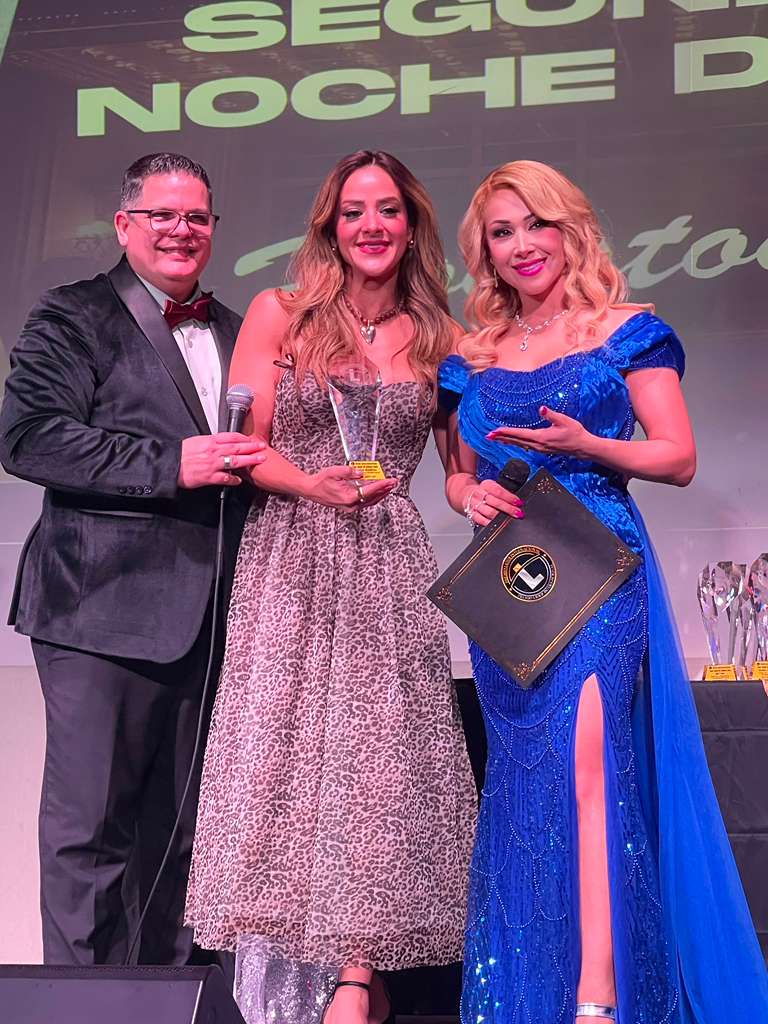 Three people in formal attire stand together on stage, holding awards, with a large screen behind them displaying text.