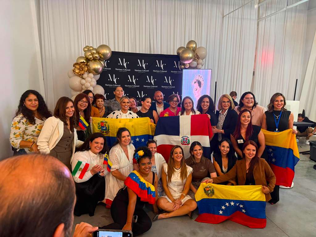 Group of people gathered indoors, holding flags and smiling, with a decorated backdrop and balloons behind them.