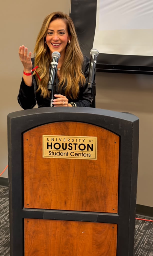 Young woman with long hair speaking at a podium, smiling, with a microphone, in a room with a screen behind her.