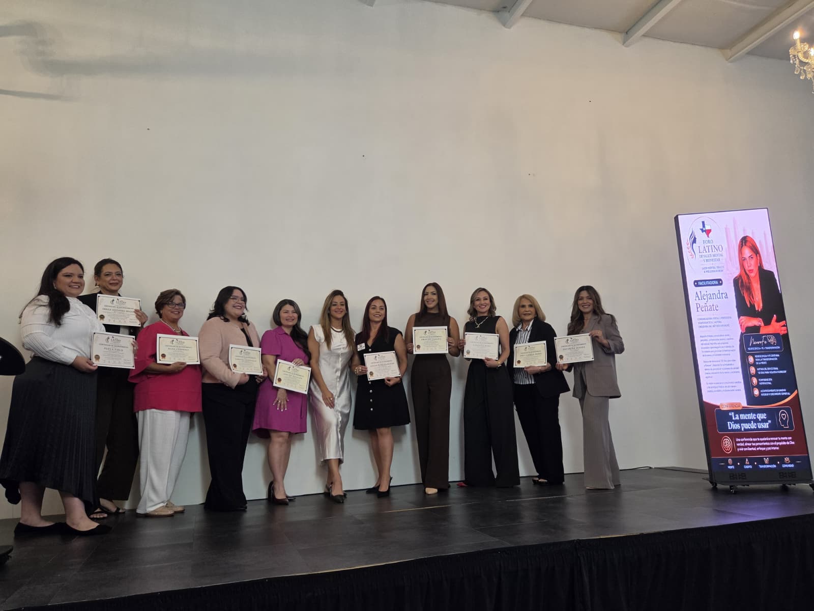 Group of women standing on stage holding certificates, with a large screen displaying a woman and text in the background.