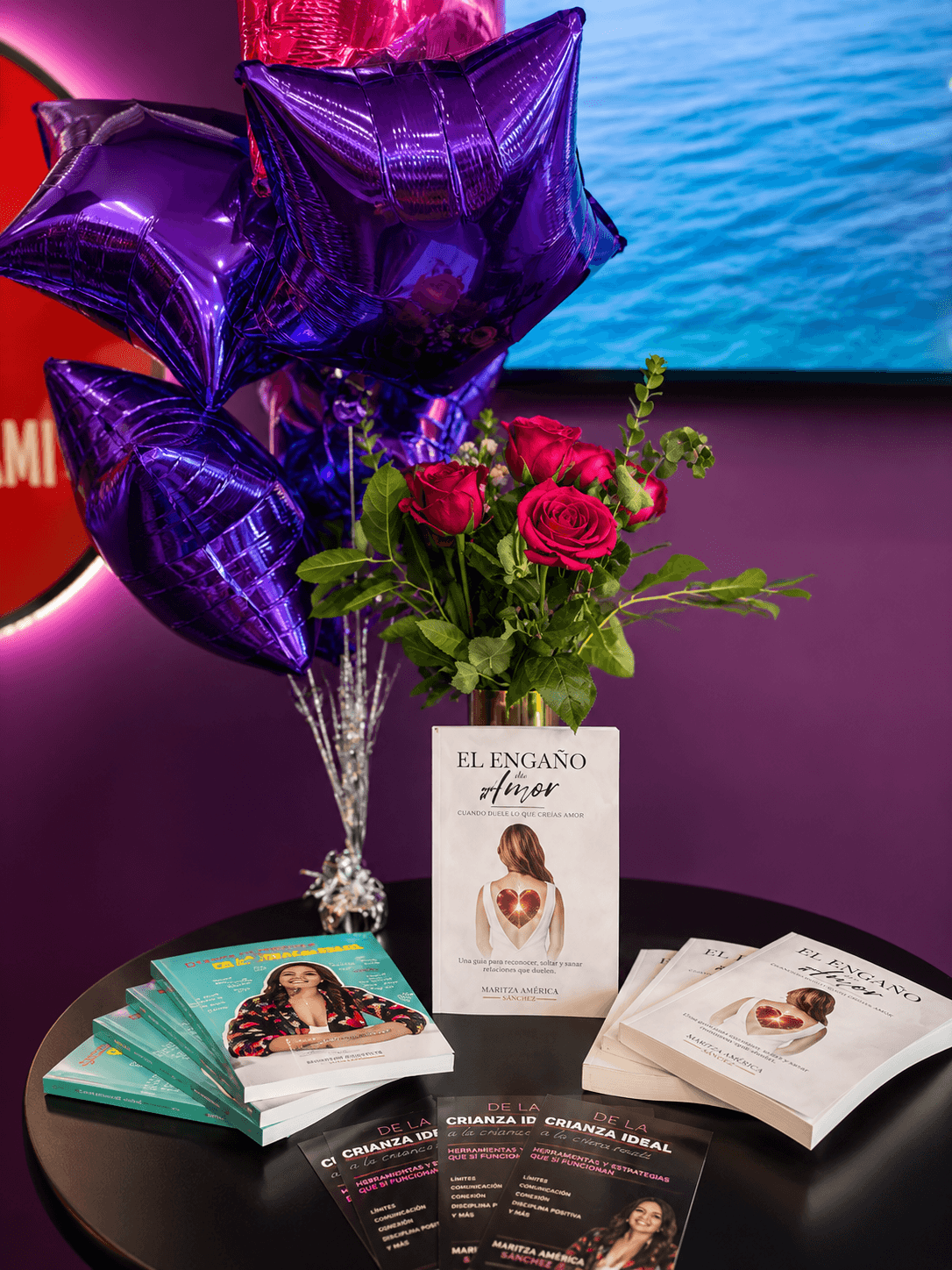 Purple star-shaped balloons, a bouquet of pink roses, and brochures on a black table against a purple and blue background.