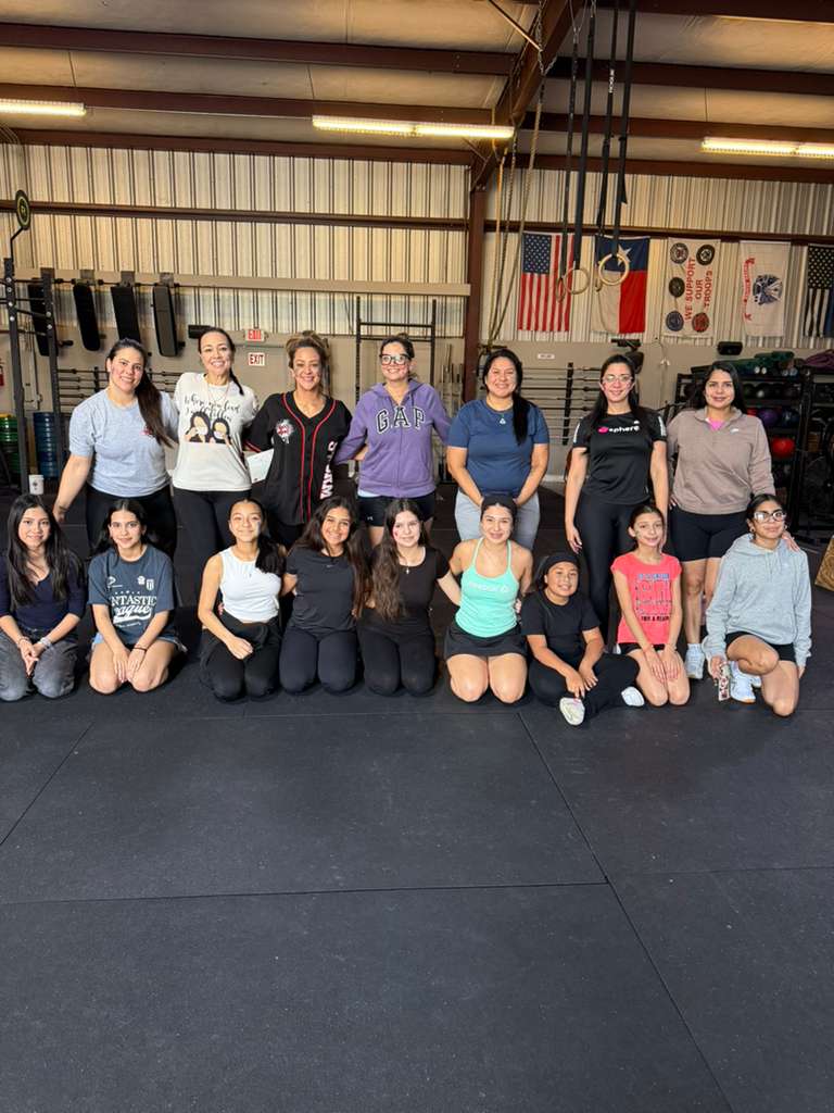 Group of fifteen women in a gym, some standing and some kneeling, with gym equipment and flags in the background.