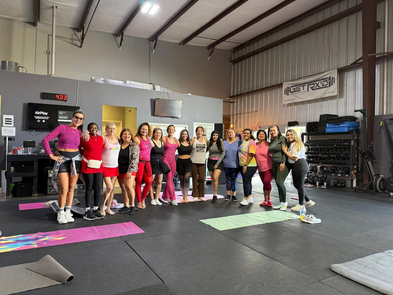 Group of women standing in a fitness studio, smiling, with exercise mats on the floor, industrial ceiling, and equipment in background.