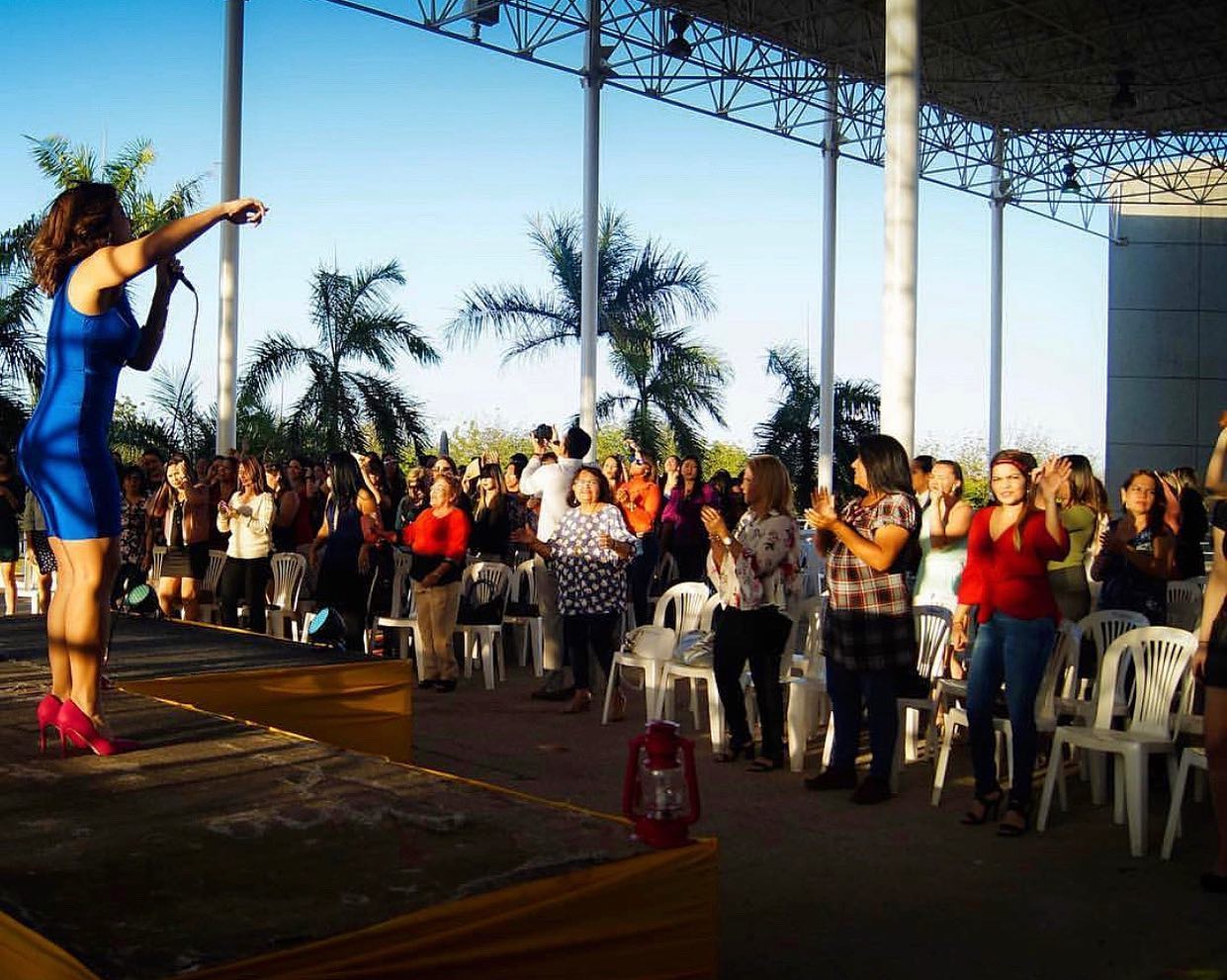 Woman in blue dress on stage pointing at audience, outdoor event with palm trees and crowd, under a large roof structure.