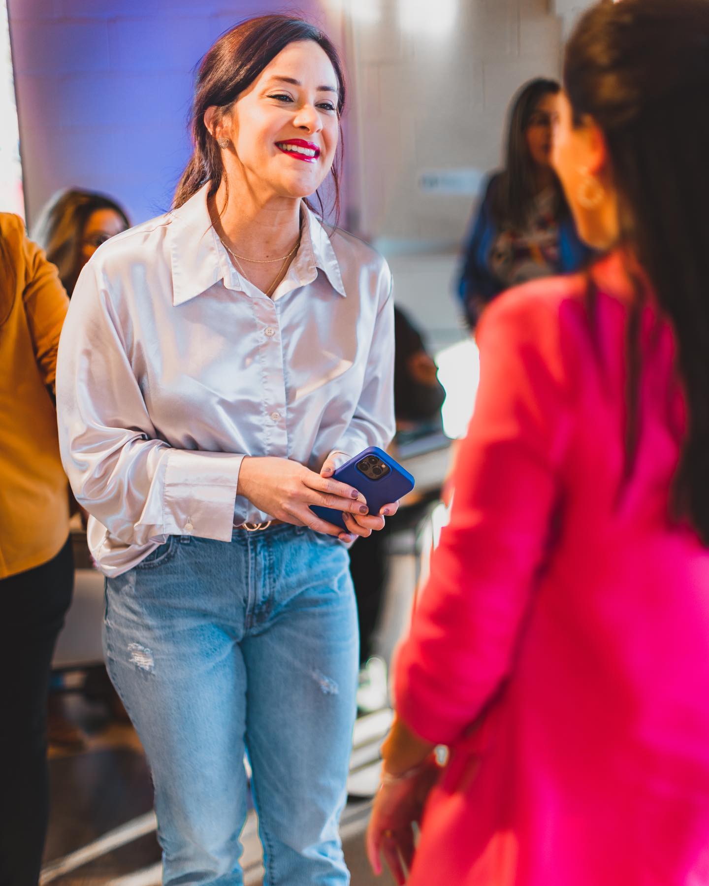 Woman smiling, holding a phone, wearing a white shirt and jeans, in a social setting with others in the background.