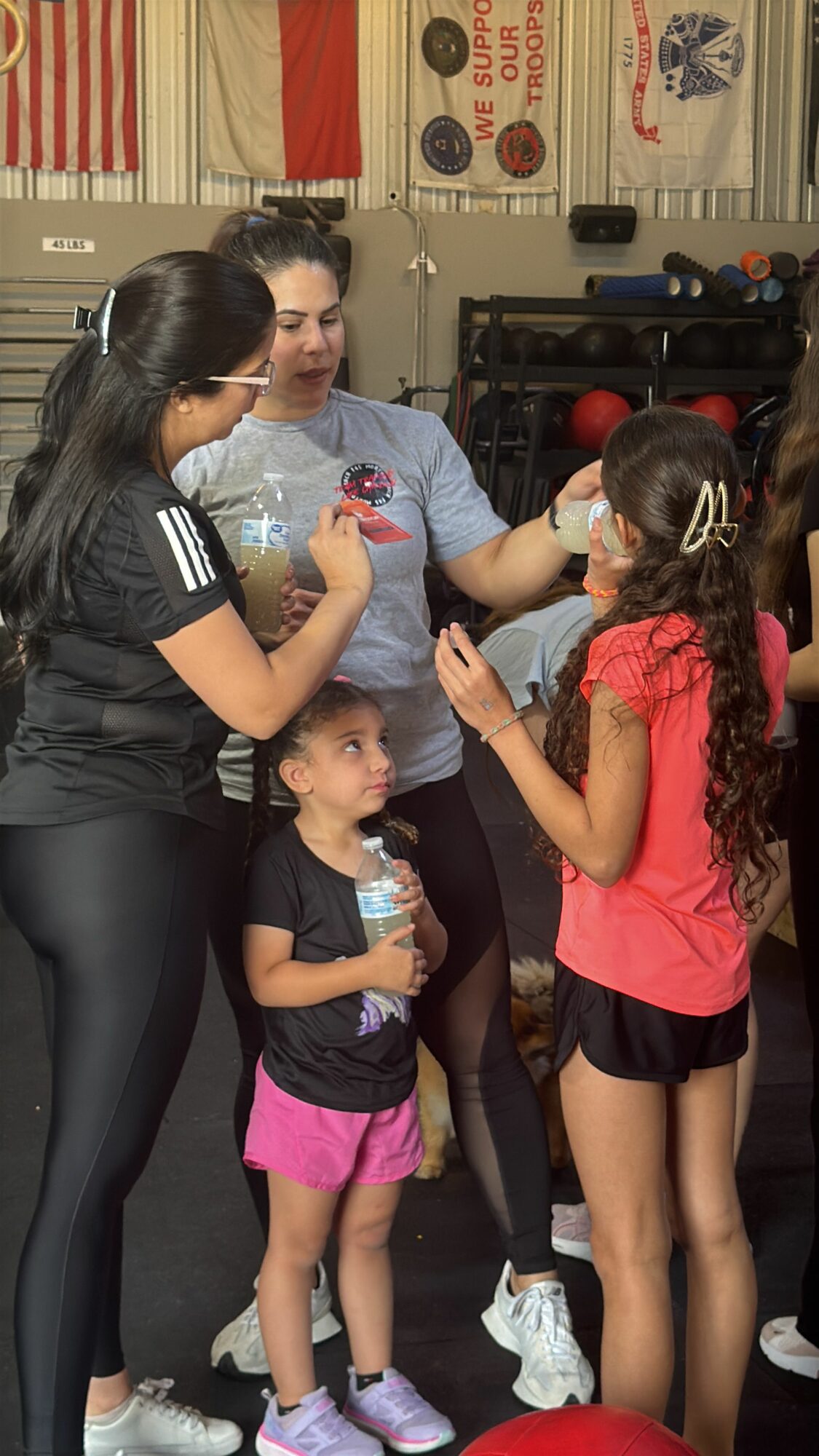 Two women and three children in a kitchen, with one woman handing a water bottle to a girl in a pink shirt. Group standing near a stove and cabinets.
