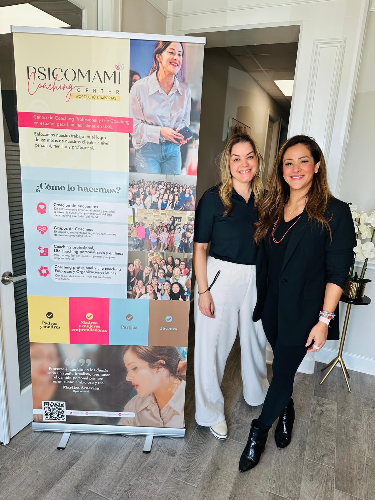 Two women standing indoors next to a vertical banner, smiling, with a white wall and door in the background.