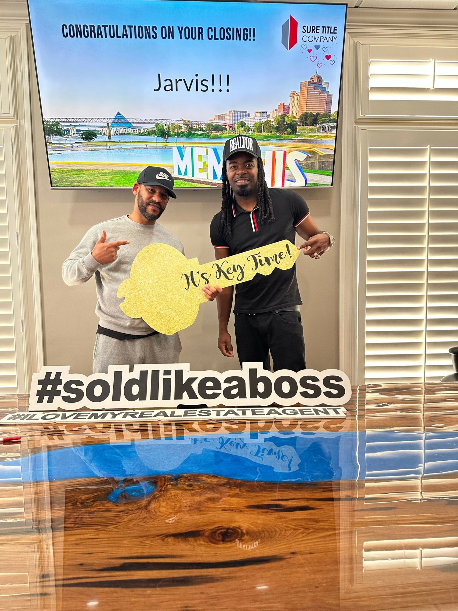 Two people holding a yellow sign with text, standing in front of a screen with a cityscape and text, on a wooden floor.