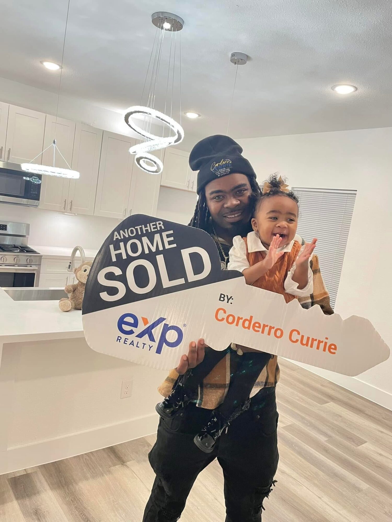 Man holding a child with a large home sold sign in a modern kitchen, smiling. Child is laughing and clapping.