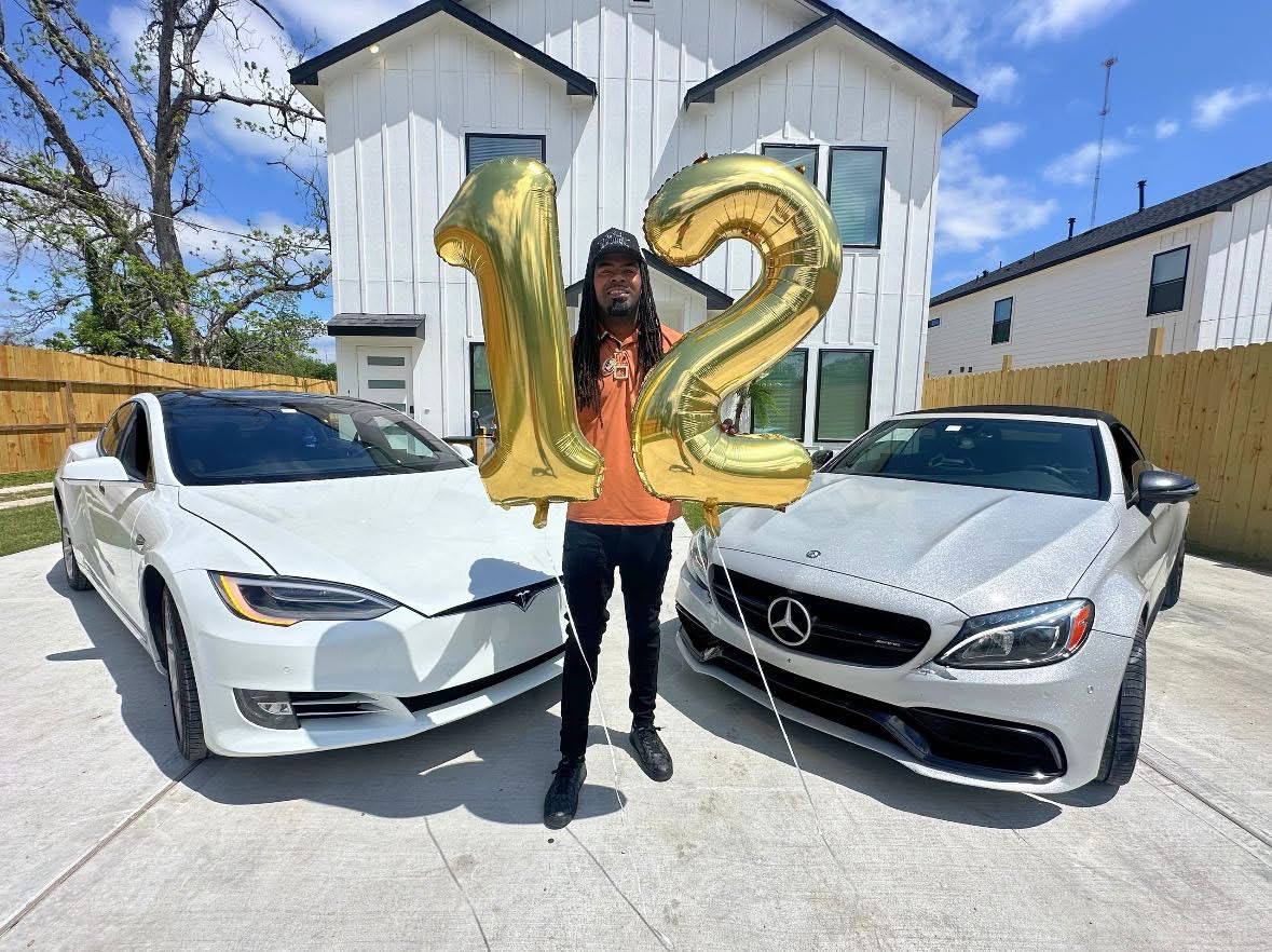 Person holding large gold balloons shaped as '1' and '2' between two silver cars in driveway, house in background.