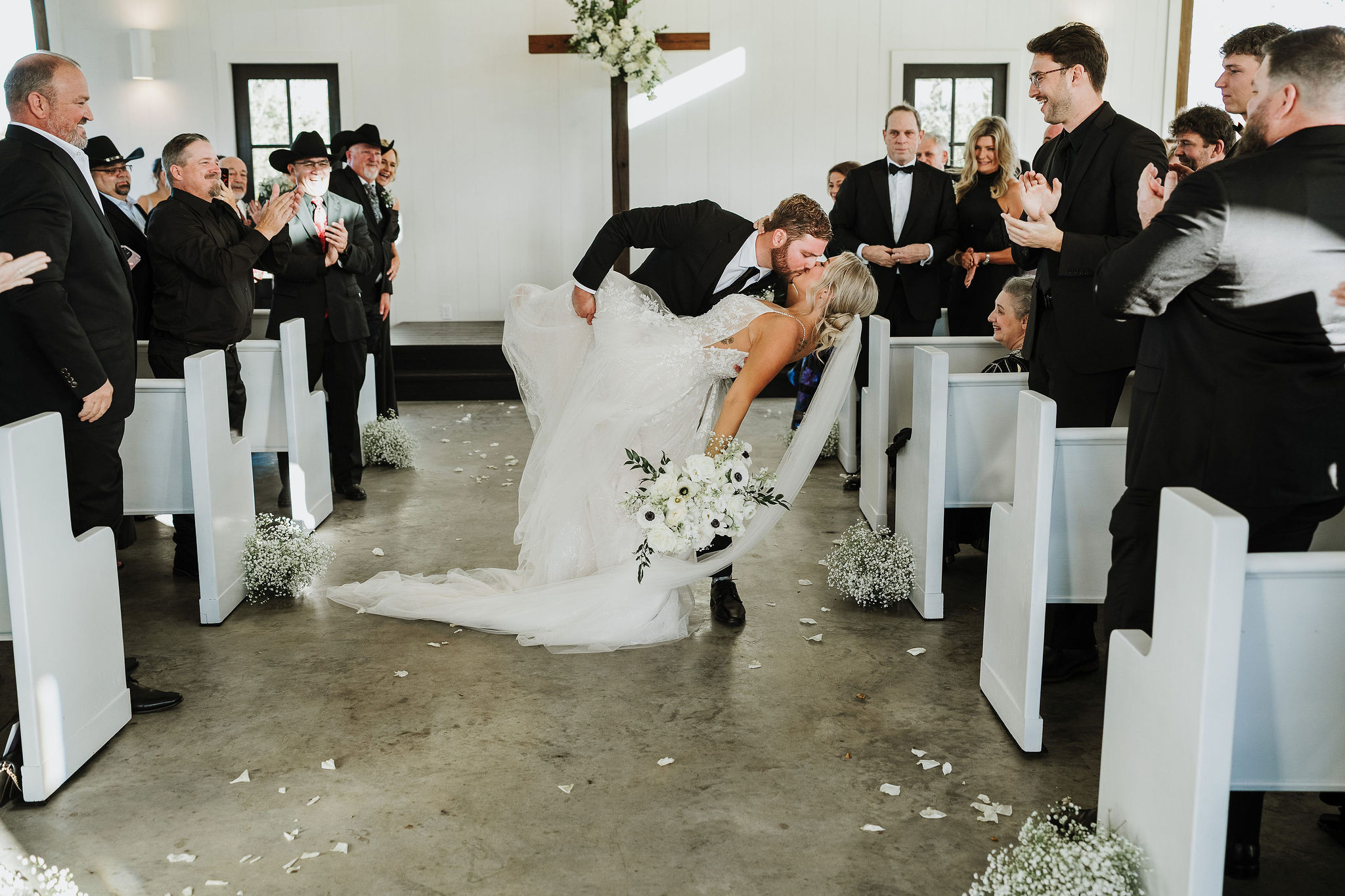 Bride and groom kiss at altar, surrounded by seated guests in a church, bride holding bouquet, groom in suit, white decor.