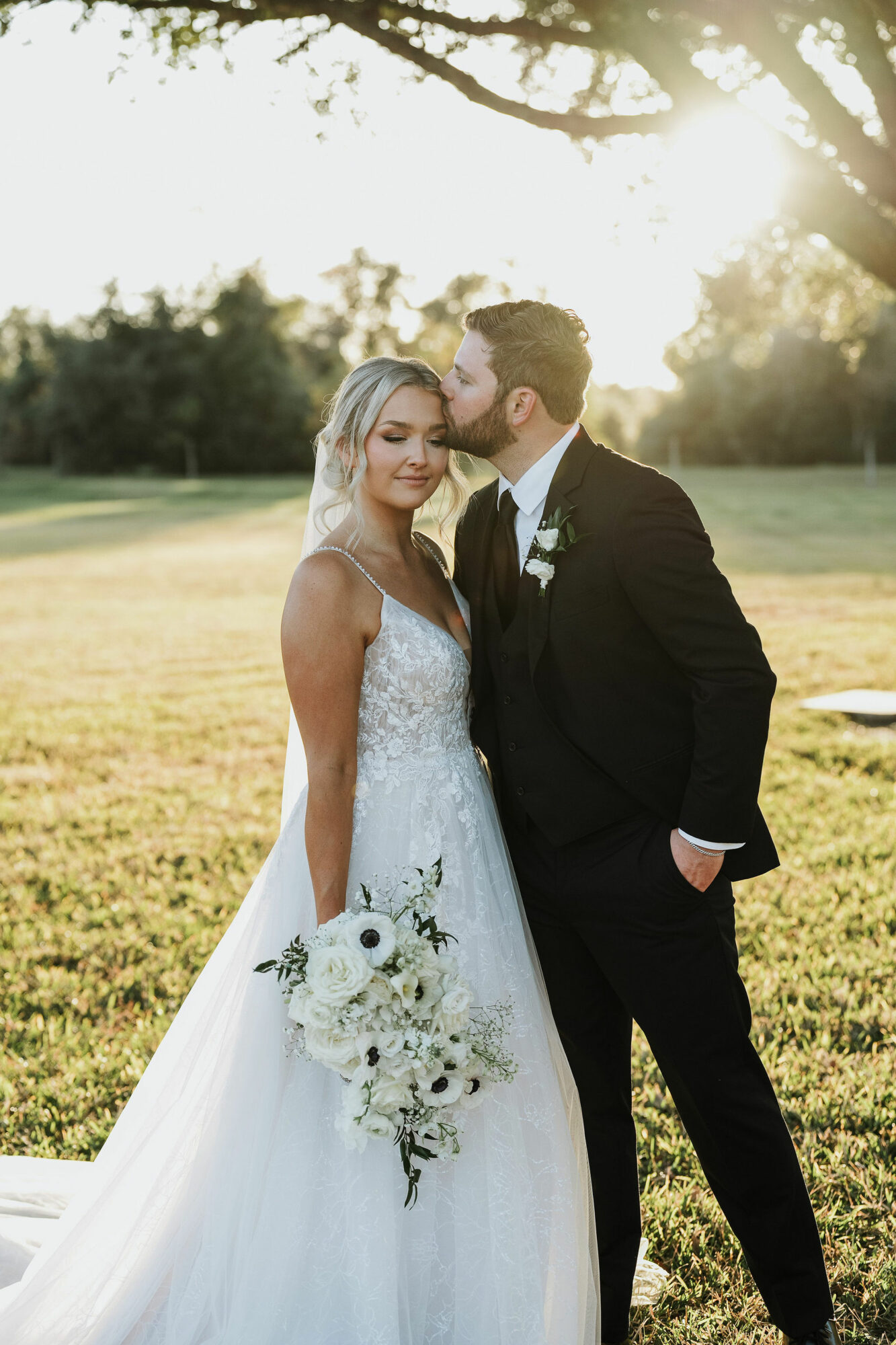 A bride and groom stand outdoors in sunlight, with trees and grass in the background, embracing each other.
