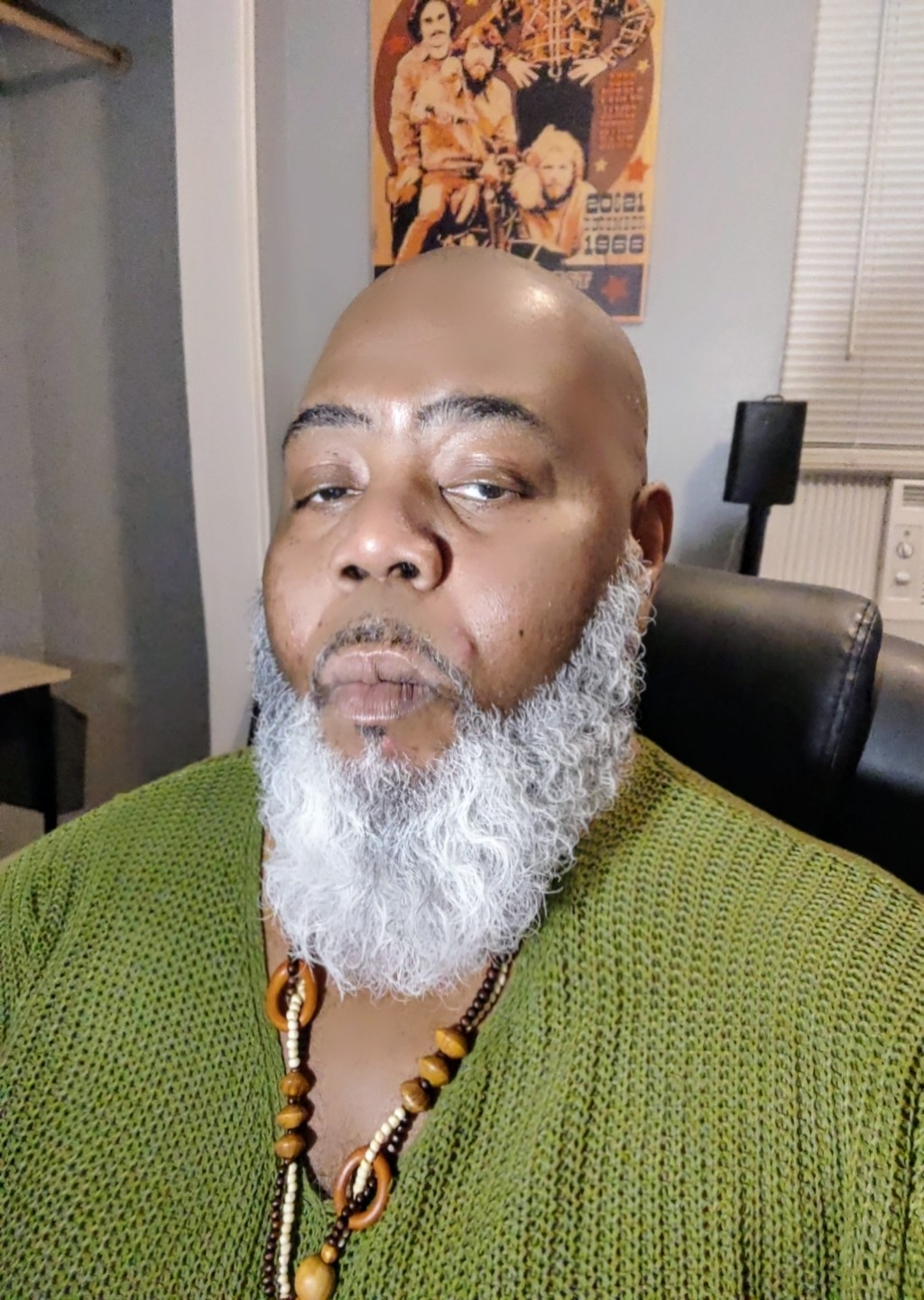 Bald man with a white beard wearing a green sweater and a beaded necklace, sitting in a room with a poster on the wall.