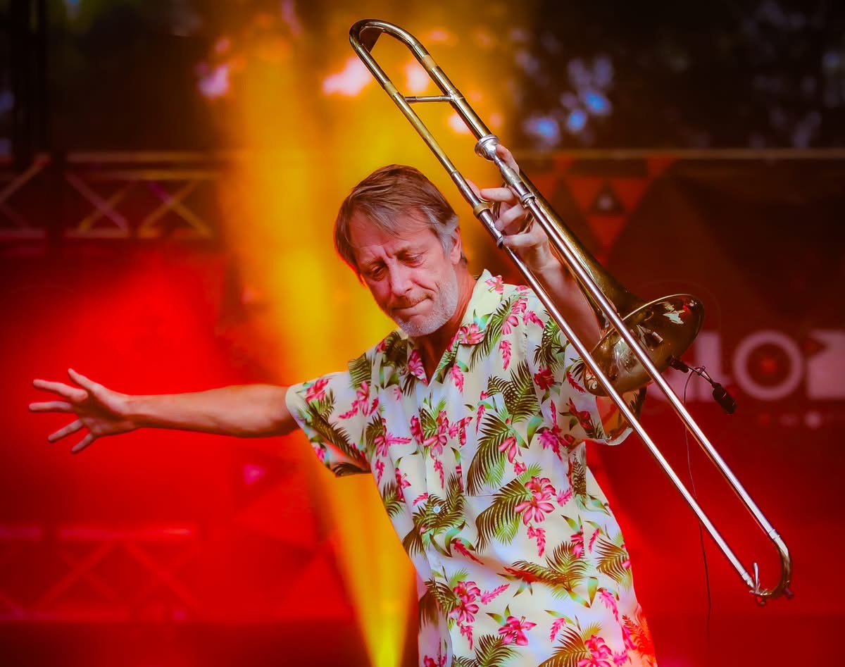 Person holding a trombone on stage with colorful lighting, wearing a floral shirt, arms extended outward.