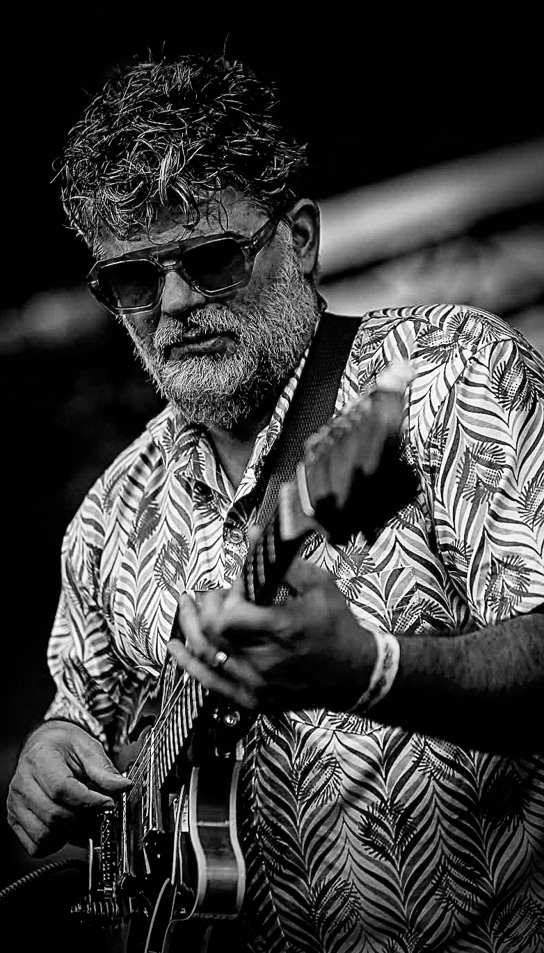 Man with curly hair and sunglasses playing an electric guitar, wearing a patterned shirt, outdoors, black and white image.