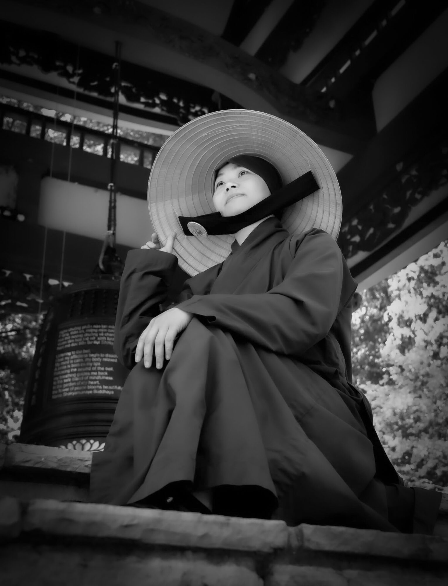 Person wearing a large hat sitting outdoors, looking upward, with trees and building structures in background.
