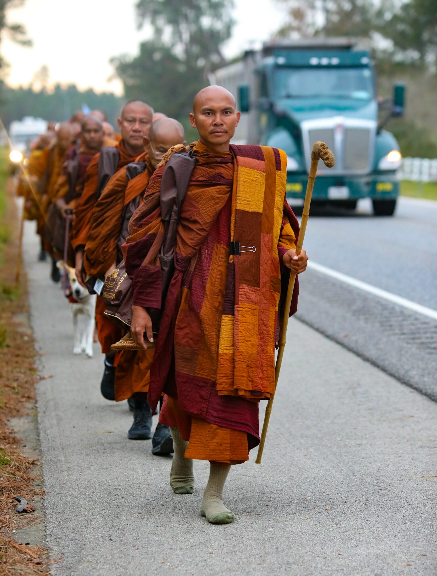 Line of Buddhist monks walking along a road, carrying staffs and wearing traditional robes, with a truck in the background.