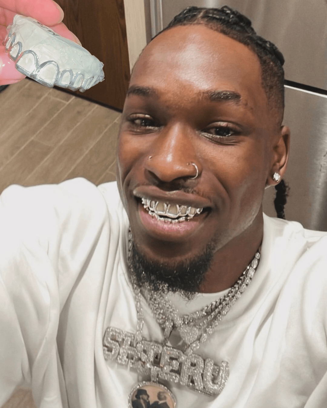 Smiling man with jewelry and a nose ring, indoors with a pink and white object in the background.