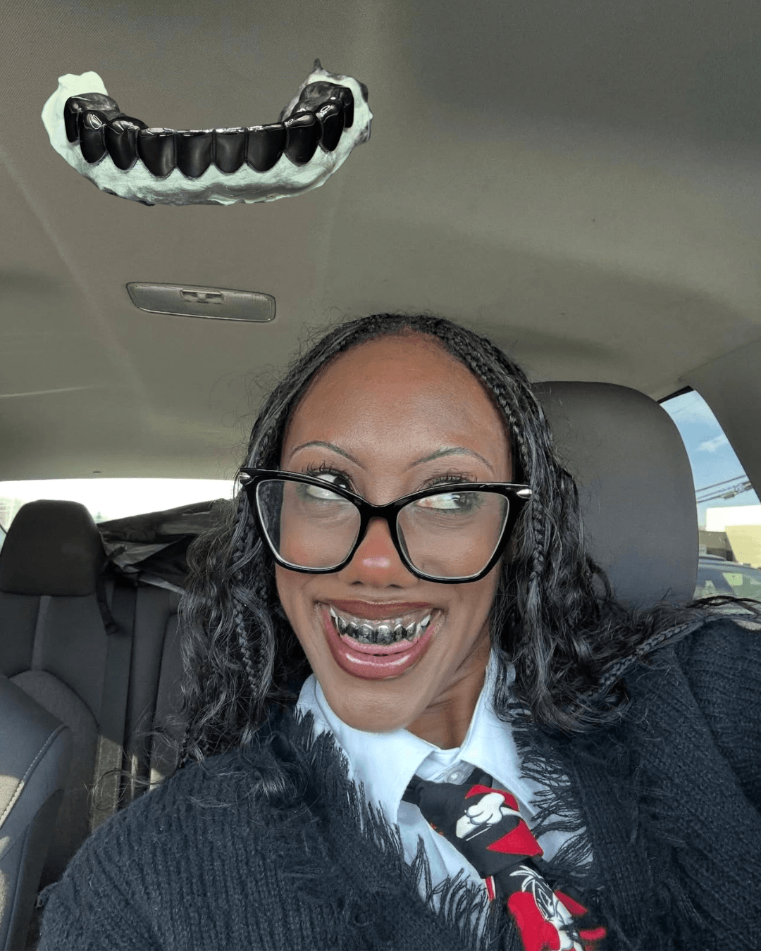 Woman with glasses smiling in car, wearing a tie and black sweater, with a dental model hanging from the ceiling.