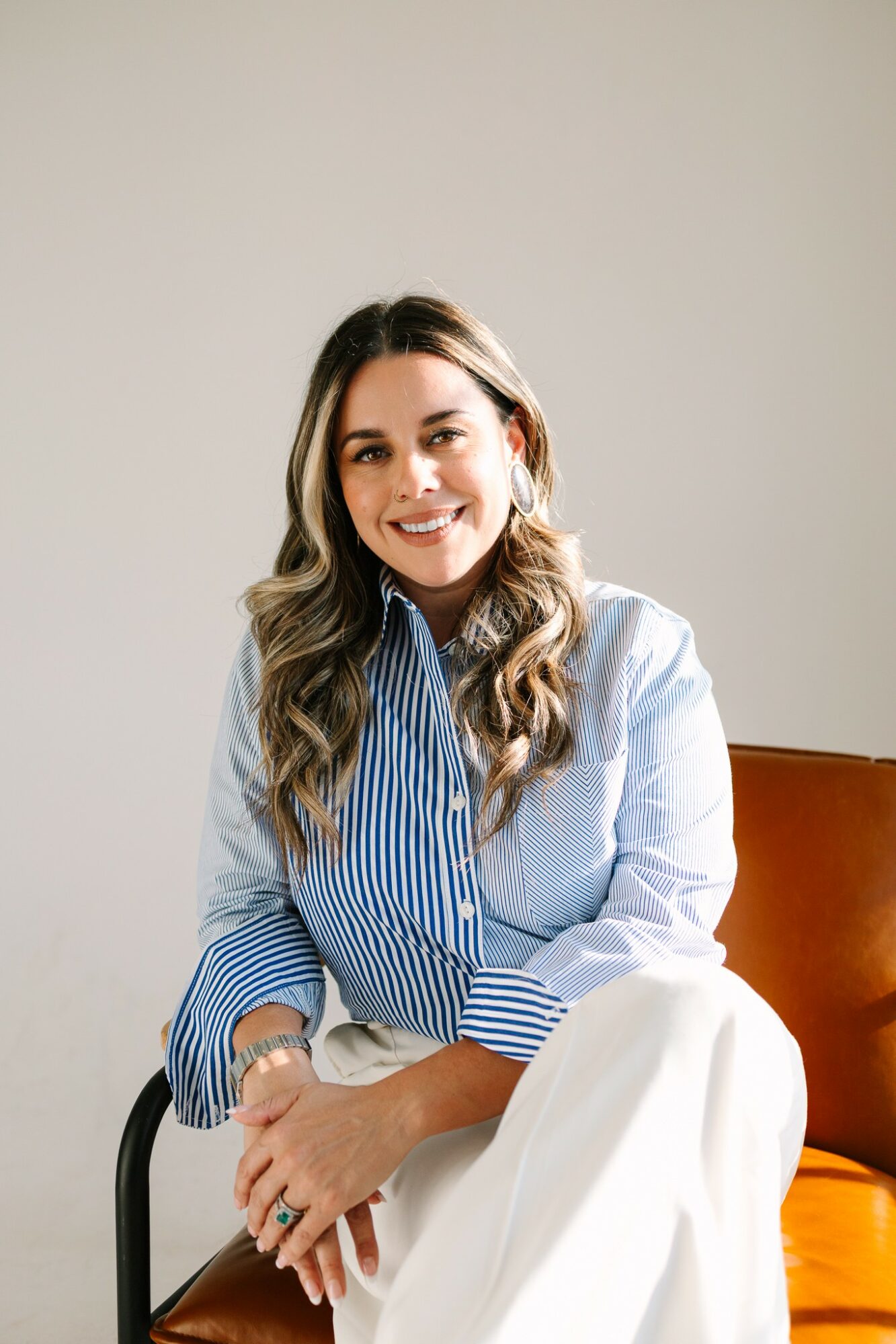 Woman with long wavy hair smiling, wearing a blue and white striped shirt, sitting on a brown chair with an orange cushion, neutral background.