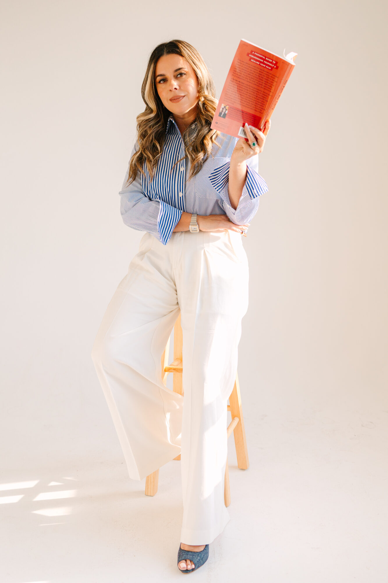 Woman with long hair in a blue striped shirt and white pants holding a red book, standing against a plain background.