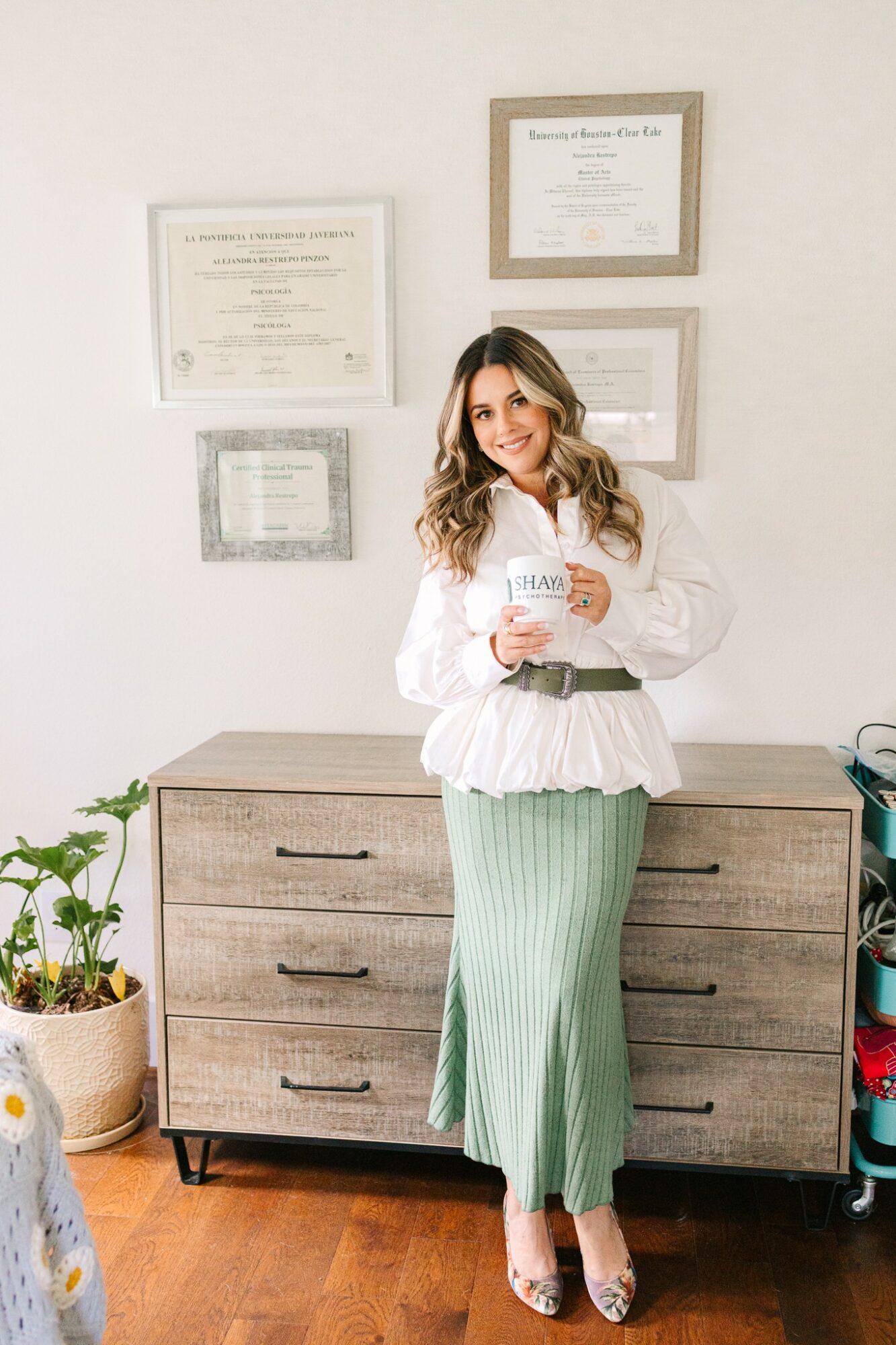 Woman standing in front of a wall with framed certificates, holding a paper, smiling, wearing a white blouse and green striped pants.
