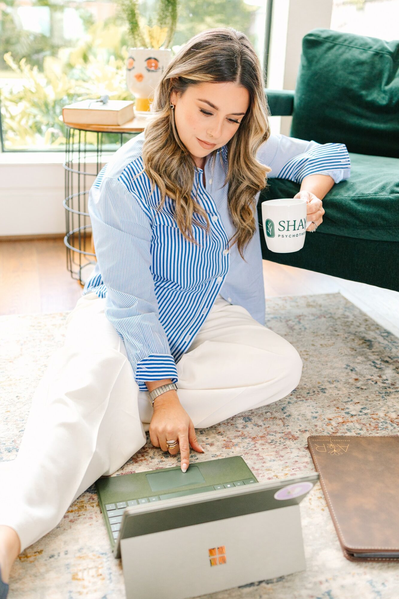 Woman sitting on floor with laptop, holding mug, in a bright room with green sofa and window.