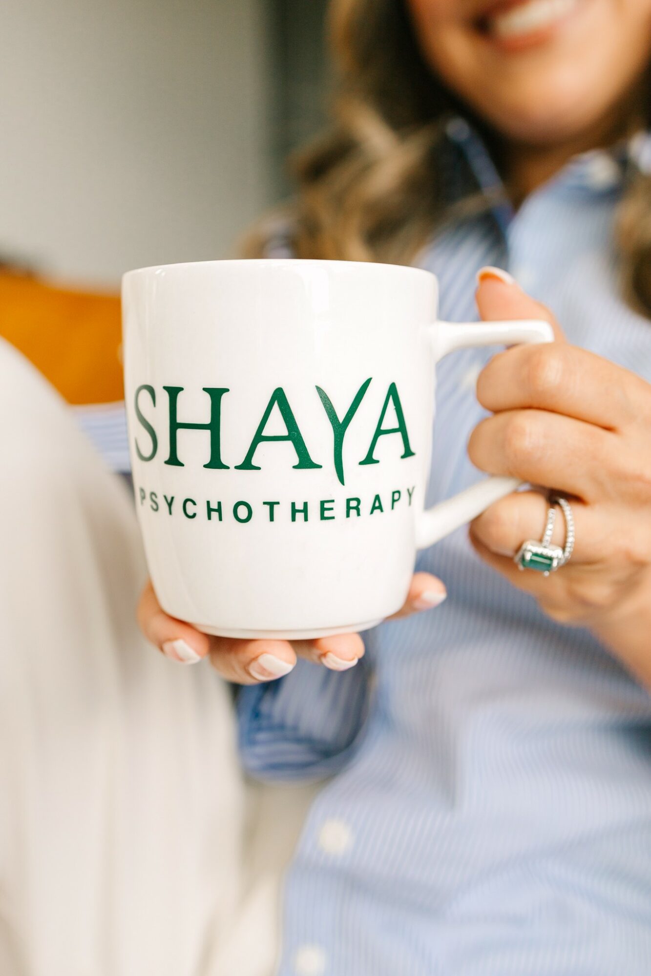 Person holding a white mug with green text reading 'SHAYA PSYCHOTHERAPY', smiling in the background.