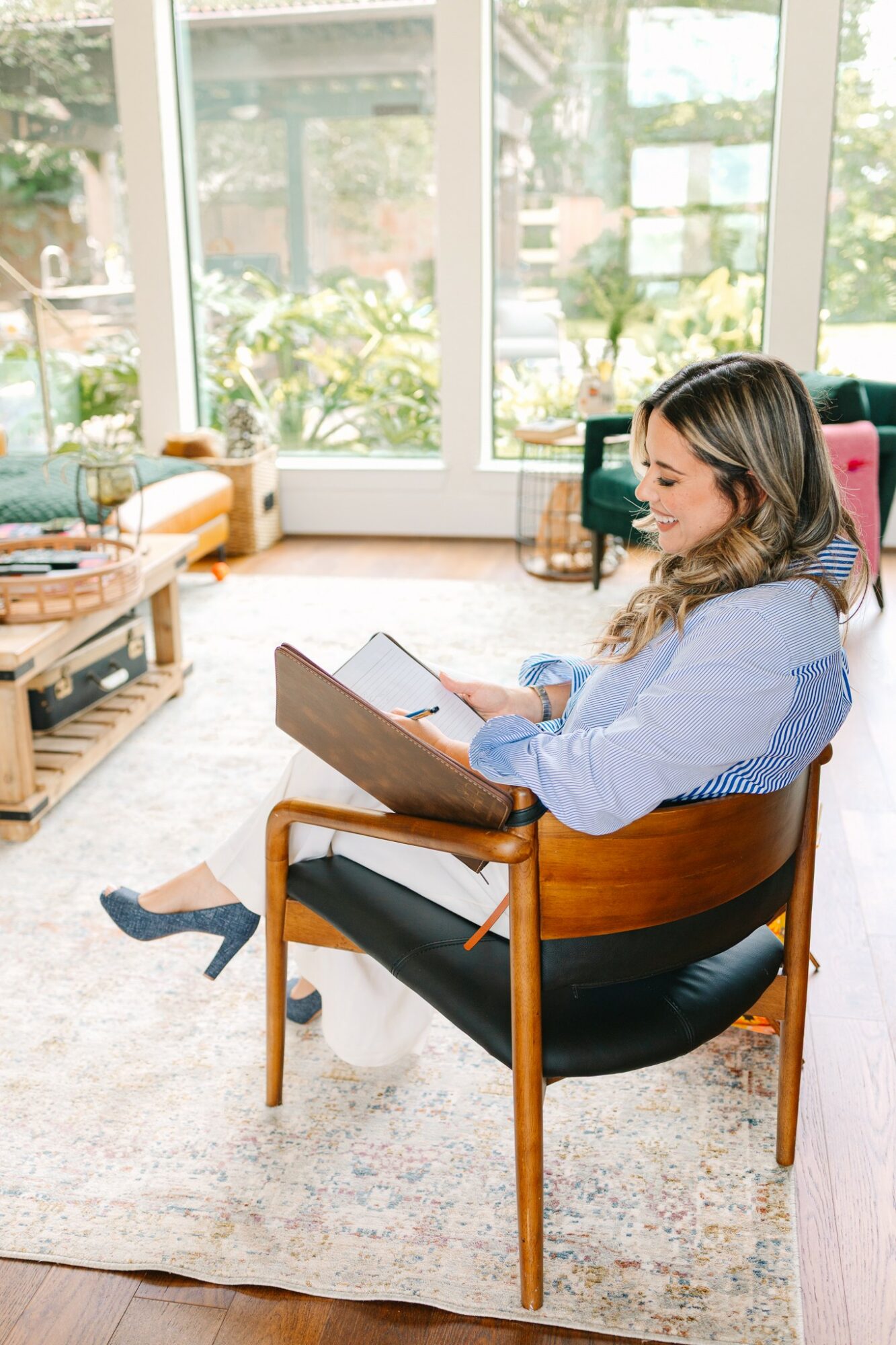 Woman sitting on a chair reading a book in a bright living room with large windows and plants.
