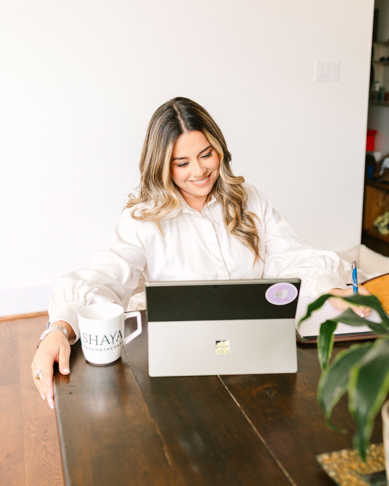Woman with long hair smiling at a tablet on a wooden table, with a mug and a plant nearby.