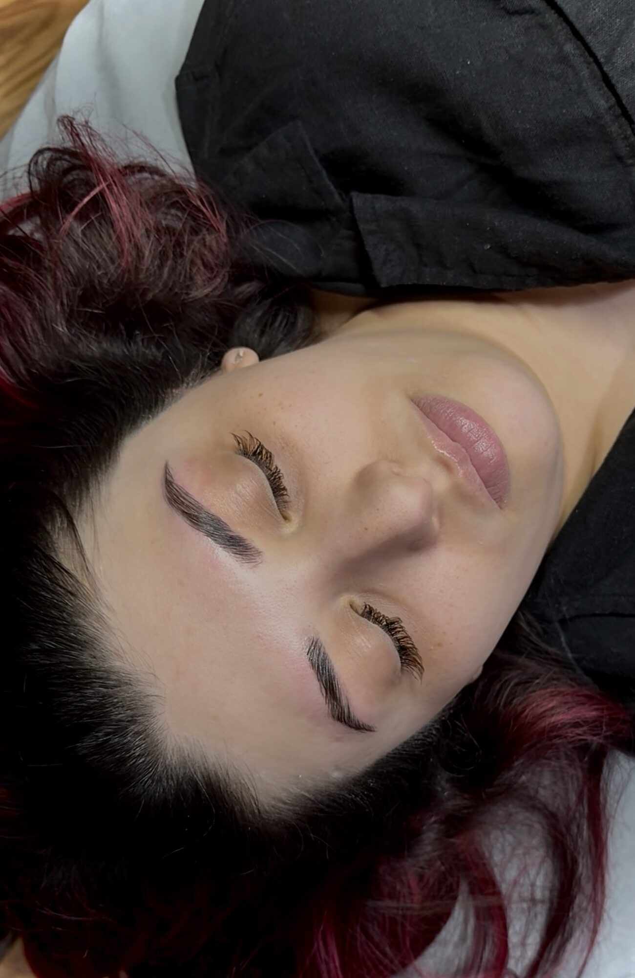 Woman with closed eyes and dark hair with red highlights, lying down, wearing a black top, relaxed expression.