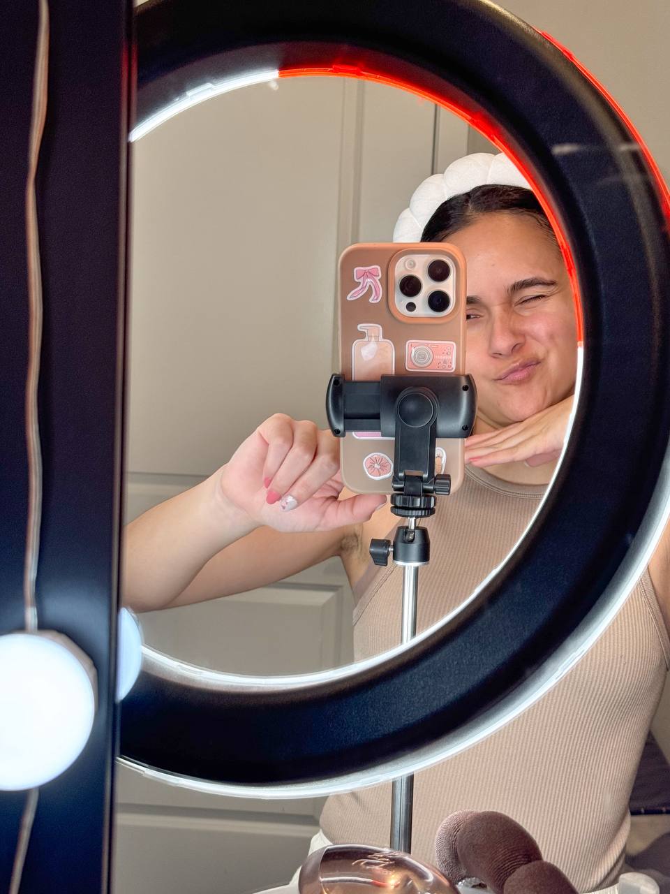 Person taking a mirror selfie with a smartphone, smiling, in a room with a ring light. The phone has a pink case with stickers.