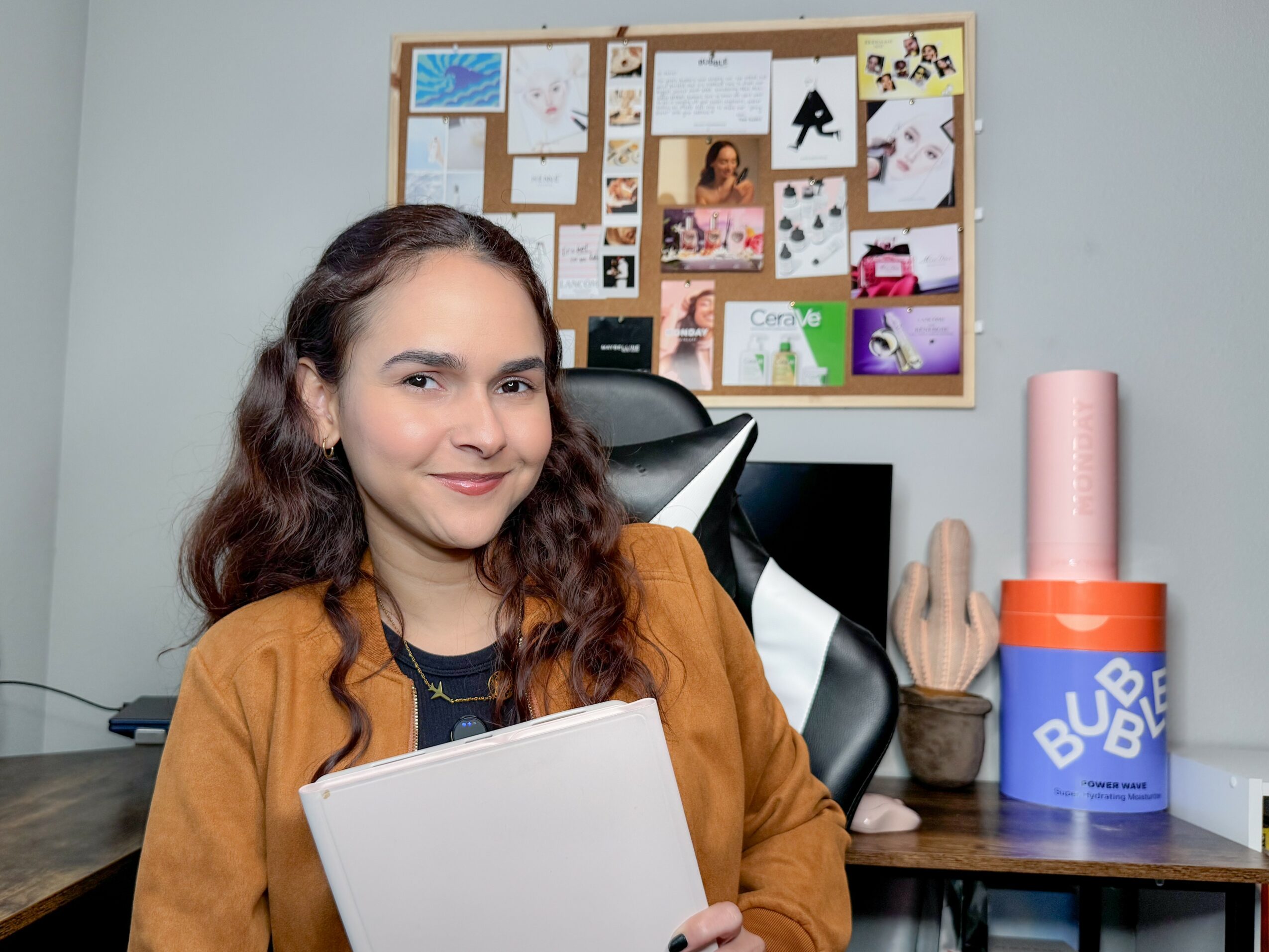 Young woman with wavy hair smiling, sitting at desk with a notebook, in front of a bulletin board, in a room.