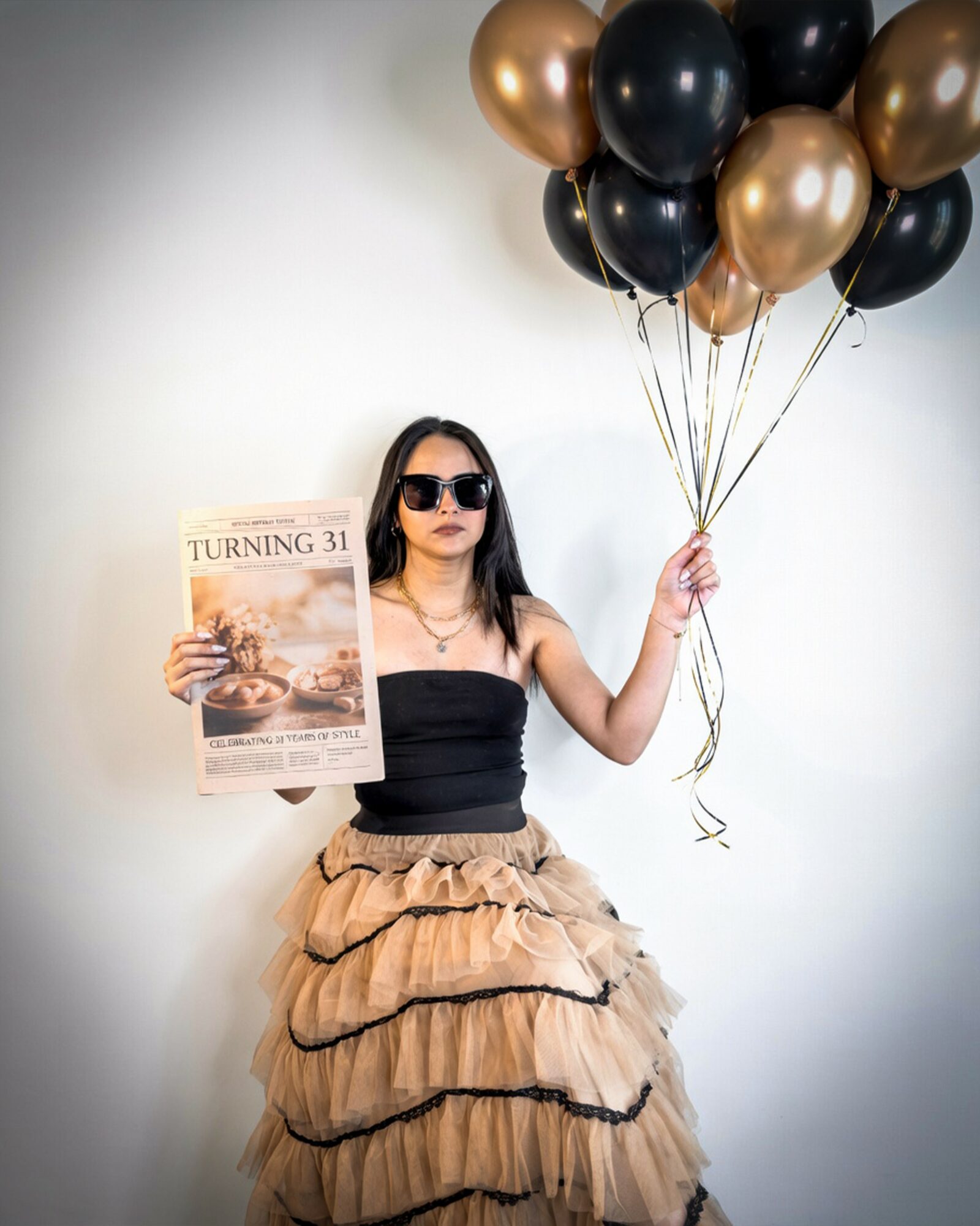 Woman holding a magazine and balloons, wearing sunglasses and a layered skirt, standing against a plain wall.
